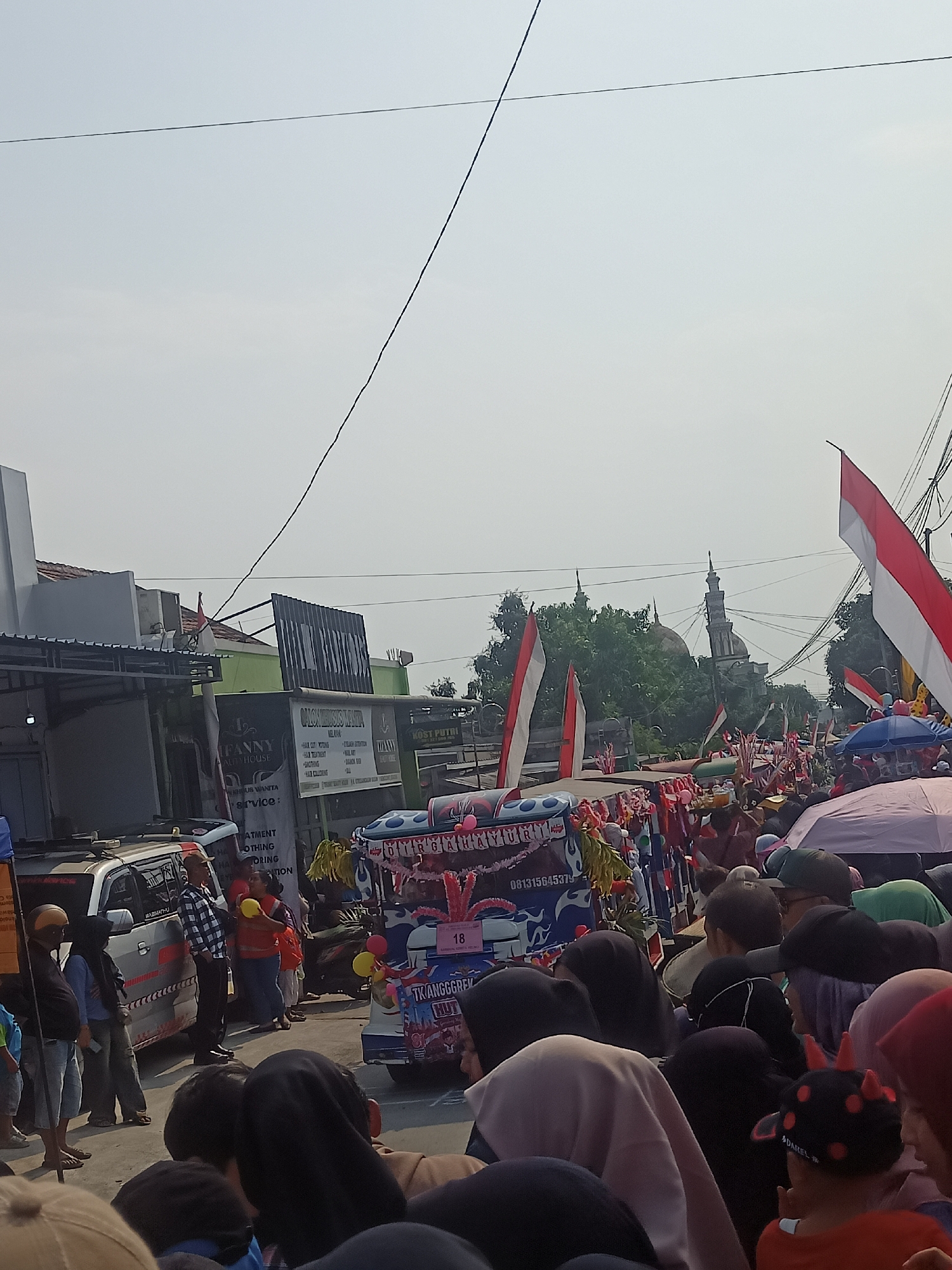 The image depicts a street scene in Gemolong, Indonesia, characterized by a procession or parade. The sky is overcast, and overhead wires are visible. A crowd of people lines the street, many of whom are looking towards the center. In the foreground, the backs of several people are visible, suggesting they are spectators. In the midground, a decorated vehicle, possibly a van or truck, is visible, adorned with flags and banners. Several Indonesian flags, red and white, are prominently displayed throughout the scene. Other vehicles, including cars, are also present. The architecture in the background suggests a built-up area with buildings and some greenery. A mosque dome and minaret are visible in the distance, indicating a religious presence in the locale. The overall atmosphere appears festive, with decorations and a gathering of people.