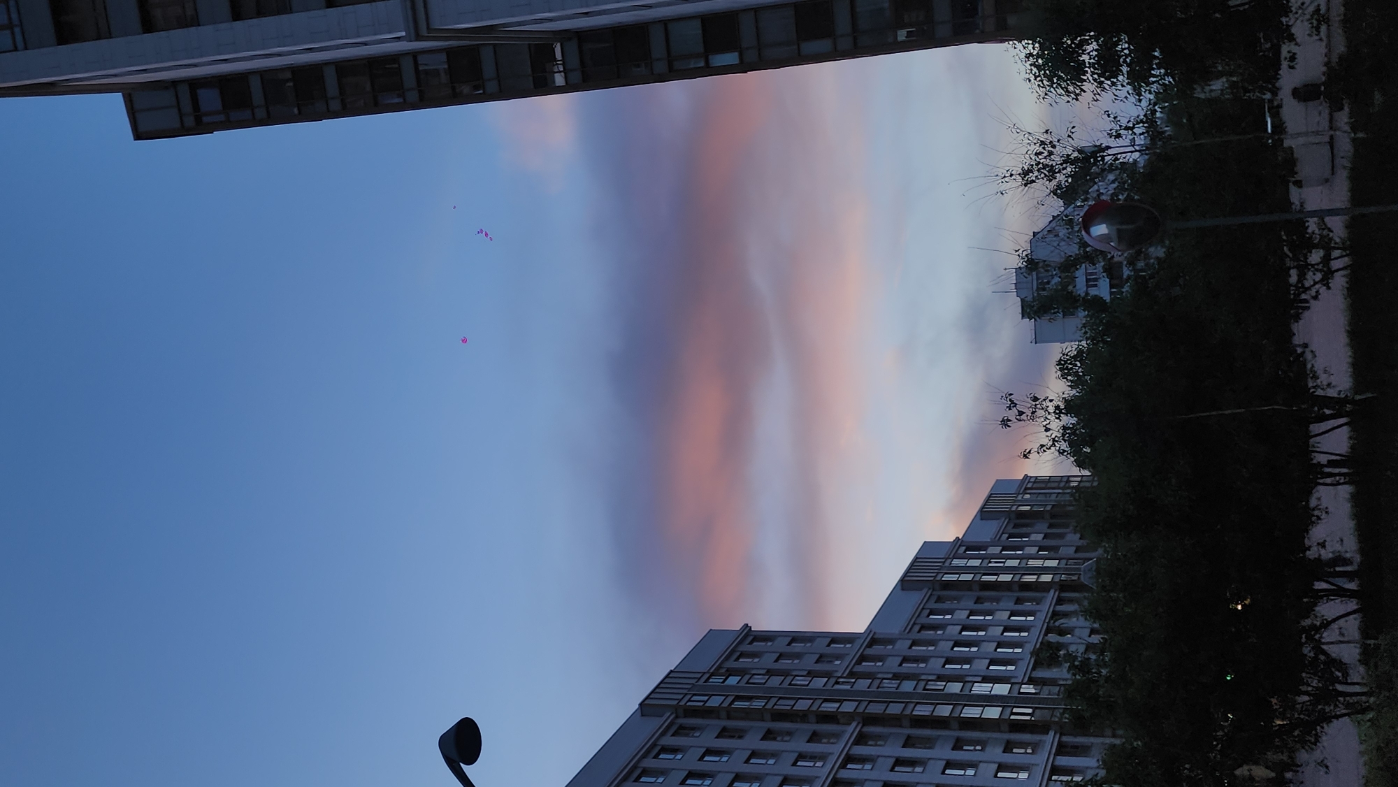 The image shows a cityscape at dusk, likely in Moscow, Russia, judging by the architecture. Two high-rise buildings flank the edges of the frame, their multiple windows suggesting residential or office use. A colorful gradient of pink, purple, and blue streaks across the sky, indicative of sunset or sunrise. In the upper part of the frame, above the left building, what appear to be balloons are visible in the sky. A street light is positioned in the lower-left corner. A street with trees are on the right of the image.