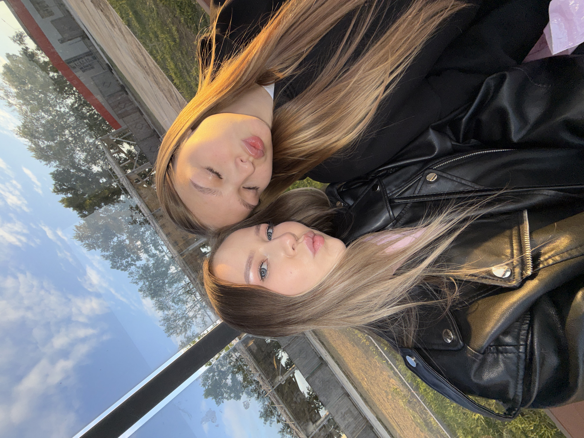 The image depicts two young women posing for a selfie in an outdoor setting, likely during the golden hour in the city of Chaikovsky, Russia. The perspective is from slightly below, looking up.
The woman on the left has long, straight, light brown hair with blonde highlights, blue eyes, and a light complexion. She is wearing a black faux leather jacket over a light-colored (possibly pink) top. Her head is angled towards the camera, and her lips are pursed in a "kissy face" gesture. Visible makeup includes light pink eyeshadow with glitter accents and lip gloss.
The woman on the right has long, straight, light brown hair, which is strongly illuminated by warm, golden sunlight on her right side. Her eyes are closed, and her head is tilted to the left, resting near the left woman. Her lips are also pursed in a "kissy face" gesture. She is wearing a dark-colored (likely black) top or jacket.
The women are positioned in close proximity, suggesting a personal connection.
The background features a prominent glass structure with a dark frame on the left, which reflects a blue sky with scattered white clouds and green deciduous trees. Beyond the immediate foreground, reflected and partially directly visible, are more trees and an industrial-style building characterized by a red upper section, grey walls, and visible scaffolding or structural elements. The ground visible is covered in green grass or dry vegetation. The scene is bathed in a warm, directional light, indicative of late afternoon or early evening sunlight.