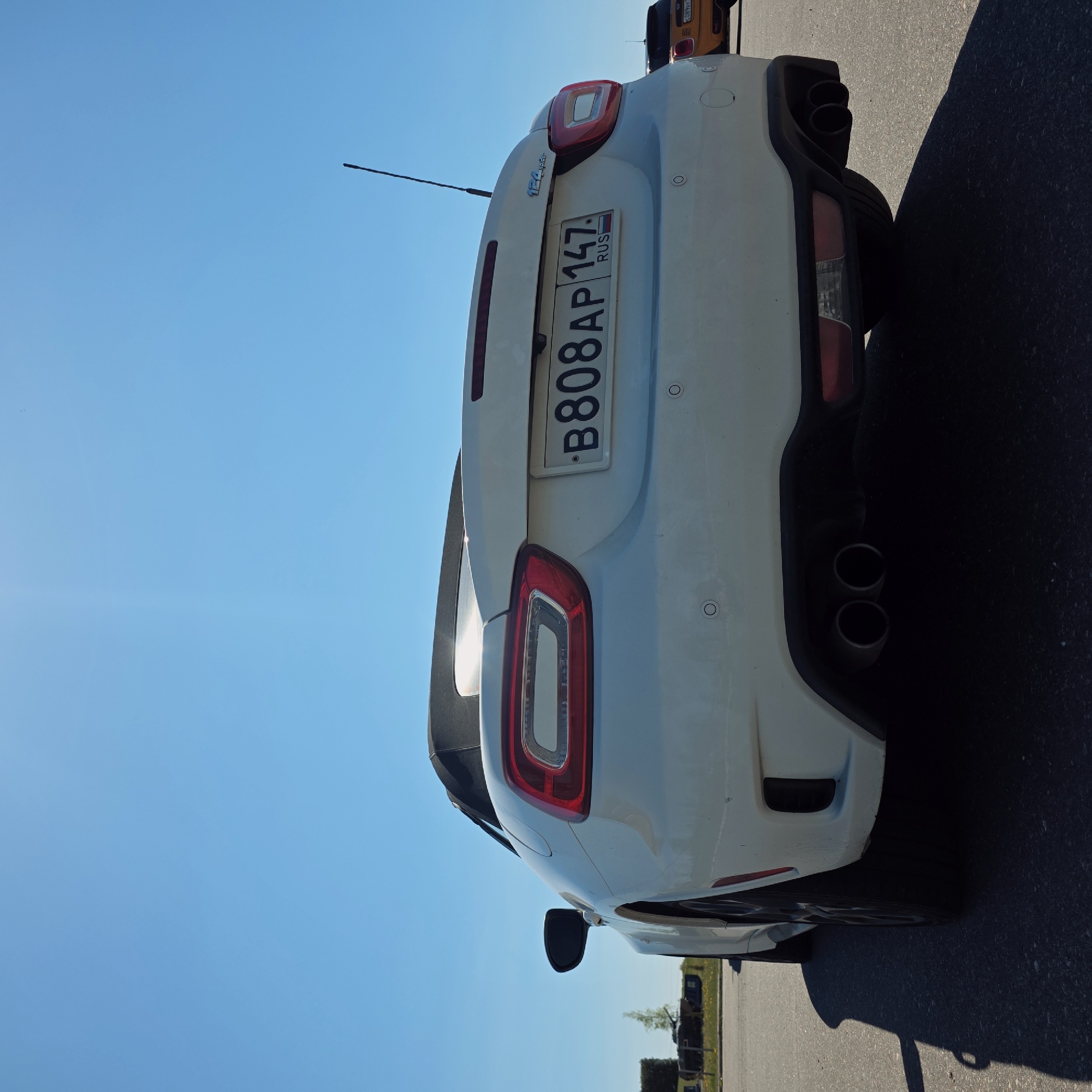 The image shows the rear of a white car with a Russian license plate "В808АР 147", a "124 Spider" emblem, and dual exhaust pipes. The car is parked on asphalt, with a bright blue sky overhead. The location is potentially Свіслацкі сельскі Савет, Belarus, given the license plate is Russian. Other vehicles are visible in the background.