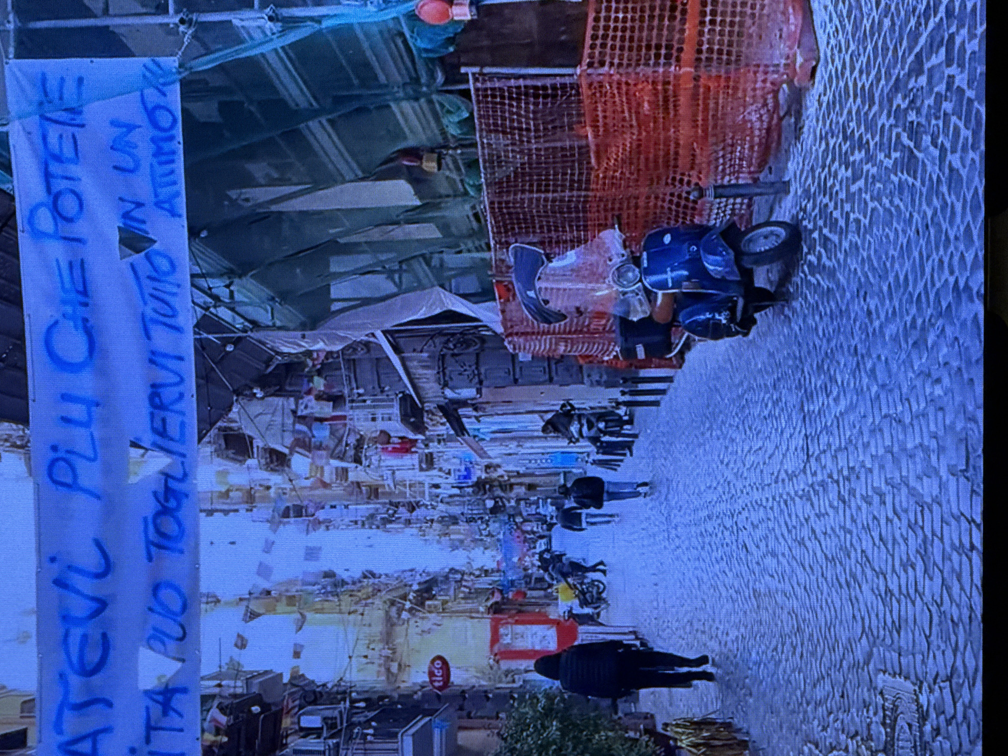 The image shows a street scene with scaffolding and construction netting in the background, suggesting an urban environment undergoing renovation or development. A blue Vespa scooter is parked on the cobblestone street. In the foreground, a white banner with blue lettering is hung vertically. The text on the banner reads: "ATEVR PIU CHE POTENTE LA VITA PUO TOGLIER TUTTO IN UN ATTIMO".
The scene appears to depict a street in an urban area, possibly a market or a busy thoroughfare, with several people walking and some parked vehicles. The lighting suggests it could be daytime. The presence of scaffolding and netting points to ongoing construction. The cobblestone street and the style of buildings in the background might indicate a European city or a historic district. The banner's message, "Life is more powerful than anything, it can take everything away in an instant," adds a reflective or cautionary tone to the scene. The overall atmosphere is one of everyday life continuing amidst construction and perhaps a moment of contemplation inspired by the banner.