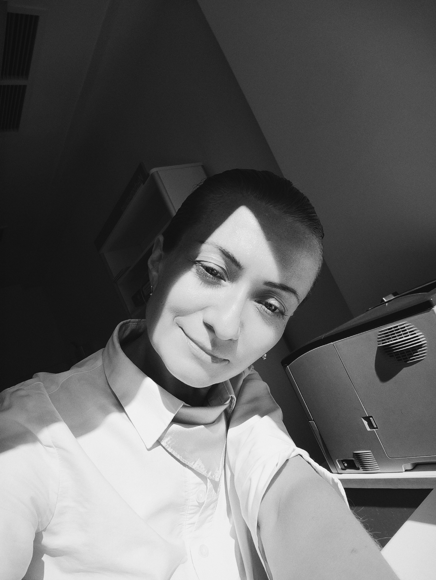 This is a black and white, high-angle, medium shot selfie of a woman. She is looking down and to her left with a slight smile. She has her hair pulled back and is wearing a collared white shirt with a dark scarf tied around her neck. Sunlight is casting shadows across her face and the room. To her right, a printer or similar office equipment is visible. The background includes a dark wall, a vent on the ceiling, and what appears to be shelving. There is no indication of the city of Moscow, Russia.