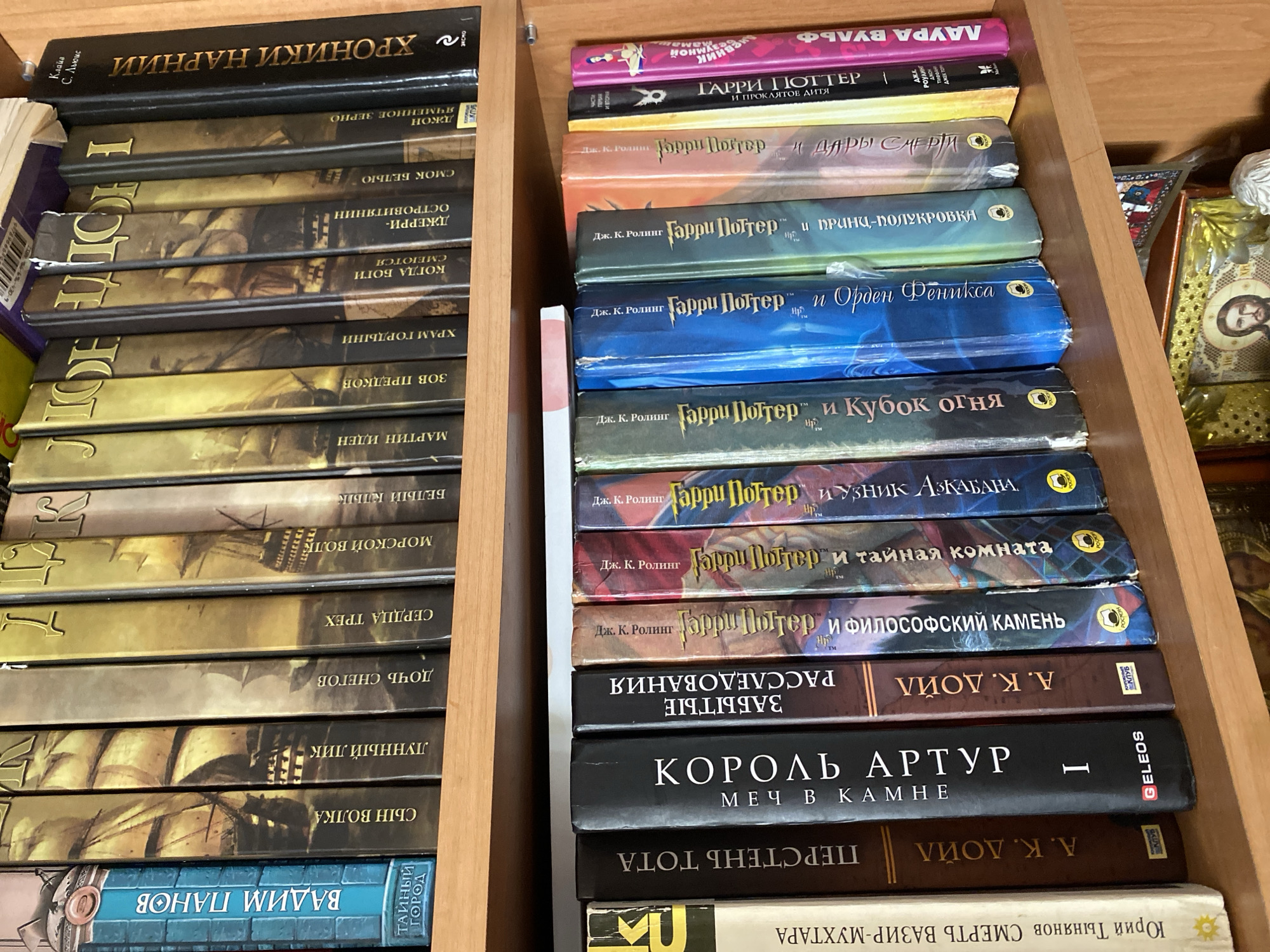 In Chornomorsk, Ukraine, two shelves of books are visible. The top shelf contains books with gold and yellow spines, featuring titles in Russian such as "Вайим Панов", "Сын Волка", "Лунный Лик", "Дочь Снегов", "Сердца Трех", "Морской Волк", "Белый Клык", "Люд", "Храм Гордыни", "Когда Бог Смеётся", "Джерри- Островитянин", "Смок Белью", "Джон Ячменное Зерно", and "Хроники Нарнии". The books are arranged vertically, with their spines facing forward. The lower shelf displays a collection of books with colorful spines, predominantly featuring the "Harry Potter" series in Russian, including titles like "Гарри Поттер и Философский Камень", "Гарри Поттер и Тайная Комната", "Гарри Поттер и Узник Азкабана", "Гарри Поттер и Кубок Огня", "Гарри Поттер и Орден Феникса", "Гарри Поттер и Принц-полукровка", "Гарри Поттер и Дары Смерти", "Гарри Поттер и Проклятое Дитя", and "Лаура Буль". Other books on this shelf include "Король Артур", "Перестрень Тота", "Забытые Рассро", and "А. К. Дойл". The books are also arranged vertically. The scene appears to be indoors, likely a home or library setting.