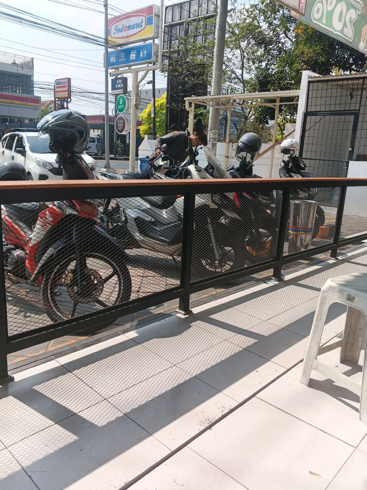 The image shows a street scene in Genuk Barat, Indonesia, with a focus on motorcycles parked in front of a building. A white car is visible in the background, along with signage for "Indomaret" and "Alfamart" convenience stores, indicating a commercial area. Several motorcycles are parked, and one has a grey helmet on its handlebars. A metal fence with a diamond pattern is in the foreground, casting shadows on the tiled ground. A white plastic stool is positioned on the right side of the frame. The sky is clear and sunny, and trees are visible in the background. The overall impression is one of a typical urban environment with retail businesses and parked vehicles.