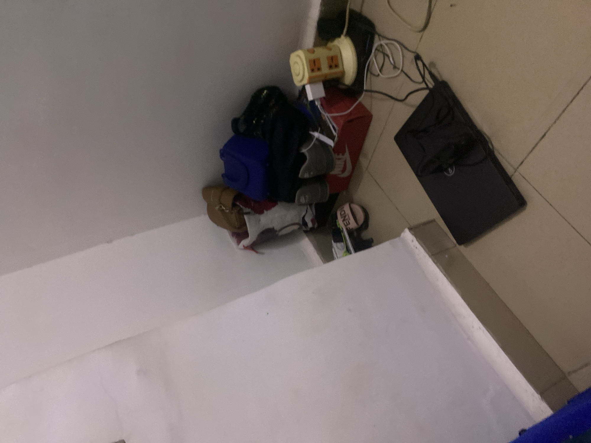 The image depicts a cluttered corner of a room, possibly a bedroom or storage area, with items piled on the floor and against the wall. In the corner, there are several bags, some appearing to be designer brands, along with shoes and a red Nike shoebox. A small, yellow electrical appliance with an orange dial sits on a black mat with several cables connected to it, including a white charging brick. A closed black laptop is placed on the tiled floor in the foreground. The walls are painted a light color, and the floor is tiled in a neutral tone. The lighting is dim, suggesting it may be evening or that the room has limited natural light. The scene does not provide any specific indicators of its location within Howayi, Nigeria, beyond being an indoor residential or storage space. There are no people or animals visible in the image.