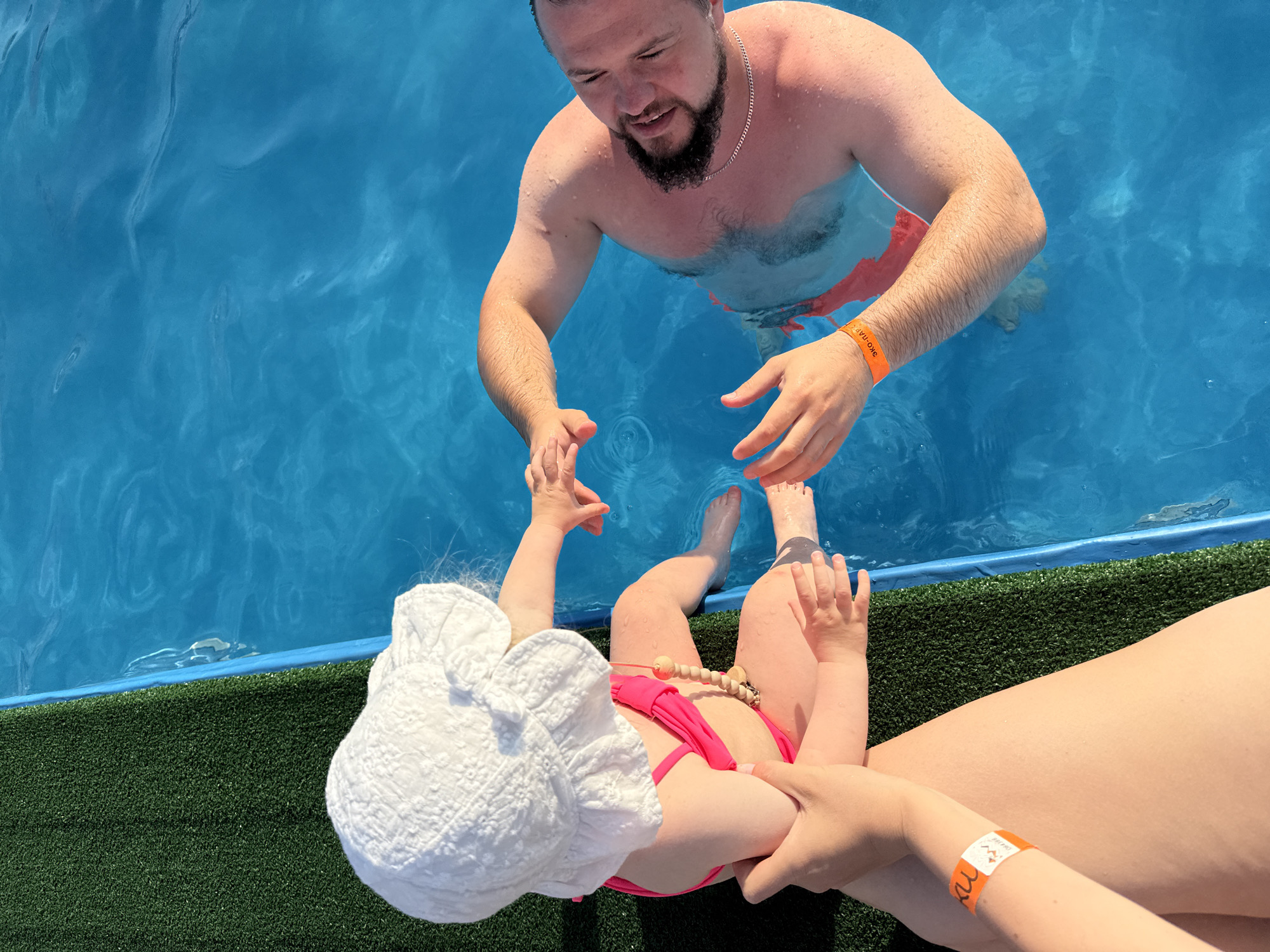In the city of Ugor, Russia, a high-angle medium shot captures a father and child at the edge of a blue swimming pool. The father, partially submerged, is on the right, facing left with a beard and wearing patterned swim trunks. He is reaching towards the child, his face showing a smile and a slightly open mouth. The child, on the left, is sitting with legs extended into the water. They are wearing a pink swimsuit and a white sun hat. The child's arms are reaching towards the father, with one hand in the water. Another person's arms are visible in the foreground, one with an orange wristband, suggesting they are holding or interacting with the child. The pool is bordered by a blue edge and a strip of artificial green turf to the left of the child. The water is clear and blue, reflecting sunlight.