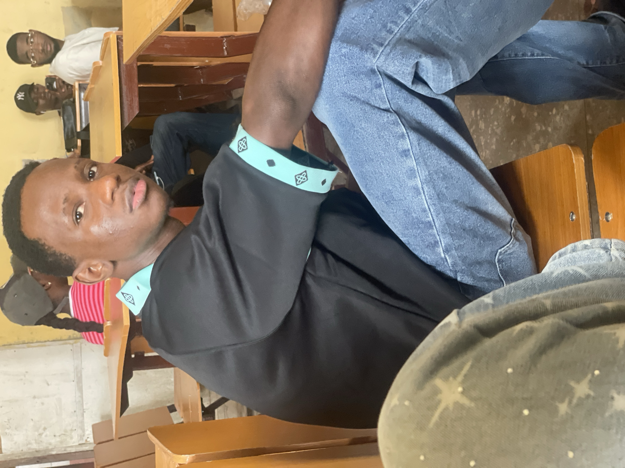 A group of young men are seated at wooden desks in a classroom in Ihiagwa, Nigeria. The primary subject, a young man in the foreground, wears a black short-sleeved shirt with a teal and black patterned trim on the collar and sleeves, and blue denim jeans. He is looking towards the camera with a neutral expression. To his left, another person is partially visible, wearing a light-colored garment with a star pattern. In the background, three other young men are seated at desks. One is wearing a grey baseball cap and a white t-shirt, holding a smartphone. Another is wearing a white t-shirt and glasses. The third is wearing a pink and white striped shirt and a grey cap with a braided ponytail. The room appears to be a classroom with simple wooden desks and chairs, and the walls are a muted yellow color. The overall atmosphere is casual and observant.