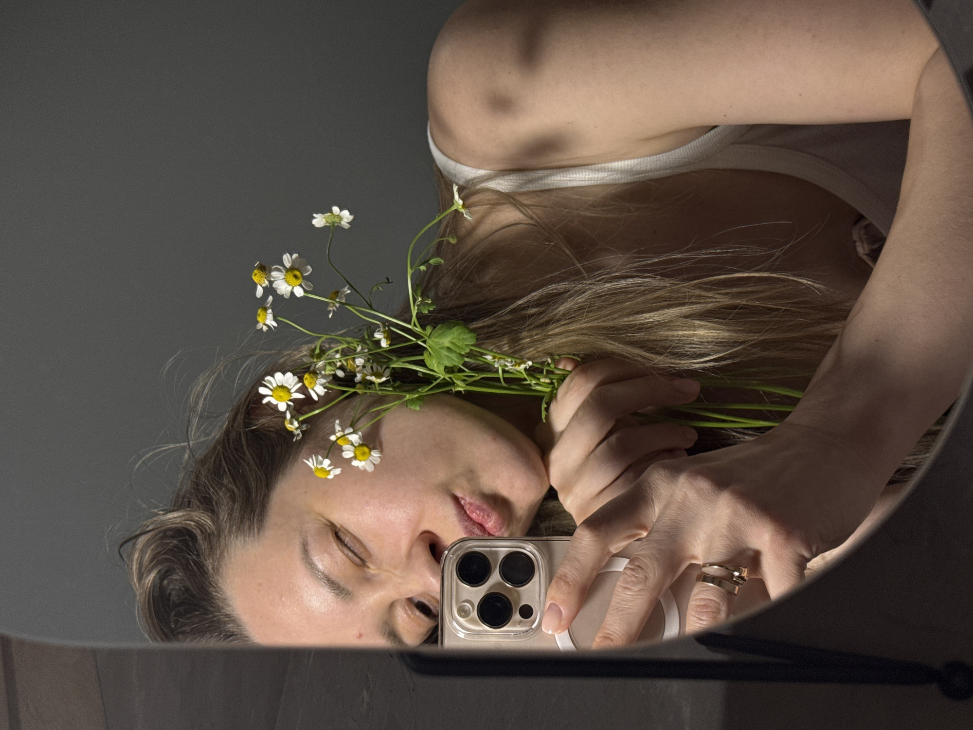 A woman is reflected in a round mirror, holding a bouquet of small daisies and a smartphone with a clear case revealing multiple camera lenses. Her eyes are closed, and she appears to be taking a selfie. She is wearing a white tank top and a double ring on her hand. The background is a solid grey color. The lower edge of the mirror is visible along with a dark object. The image was taken in a building, likely inside the city of Chaikovsky, Russia.