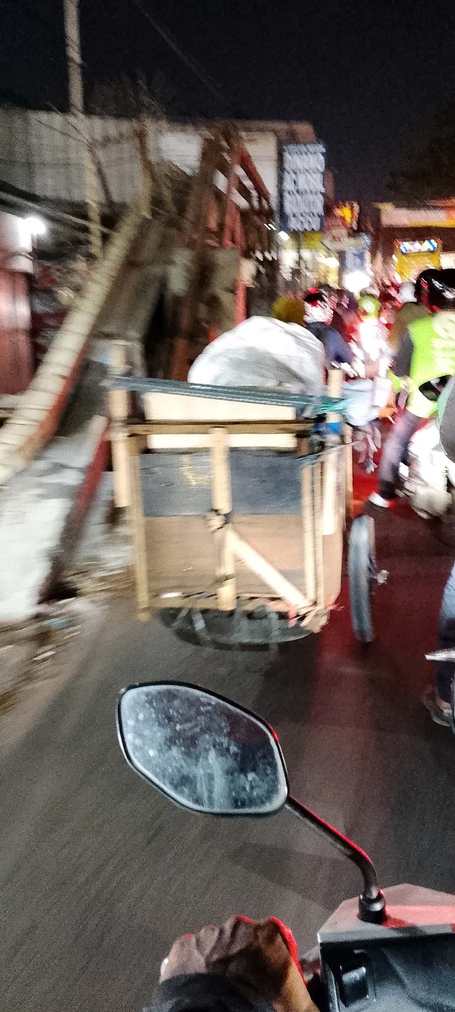 The image depicts a street scene at night in Gunung Putri, Indonesia. The foreground shows the perspective of someone riding a motorcycle, with a rearview mirror and a hand on the handlebar visible. In the mid-ground, a wooden cart with metal reinforcements is being pulled or pushed. The cart contains a large, white, tarp-covered object. The background is blurred but suggests a busy street with other motorcycles, some carrying passengers, and illuminated signs. There appears to be some construction or debris in the background as well. The overall lighting is dim, characteristic of nighttime.