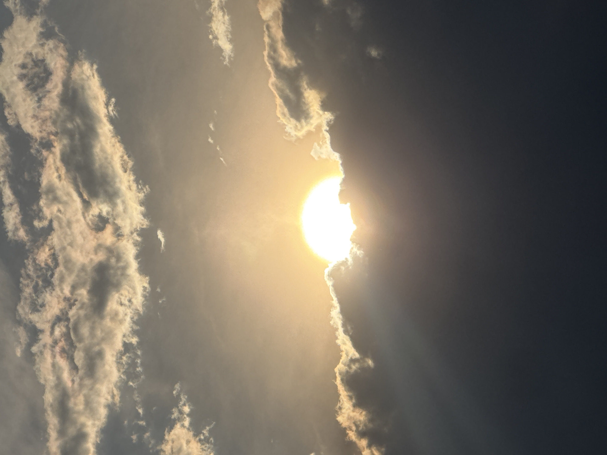 The image depicts a bright sun partially obscured by clouds. The clouds are various shades of gray and white, with the sun's light shining through the gaps. The sky transitions from a bright, sunlit area near the sun to a darker shade on the right side of the image. The image appears to be taken from a low angle, looking up at the sky. The location in San Francisco Ocotlán, Mexico is not discernible from the image.