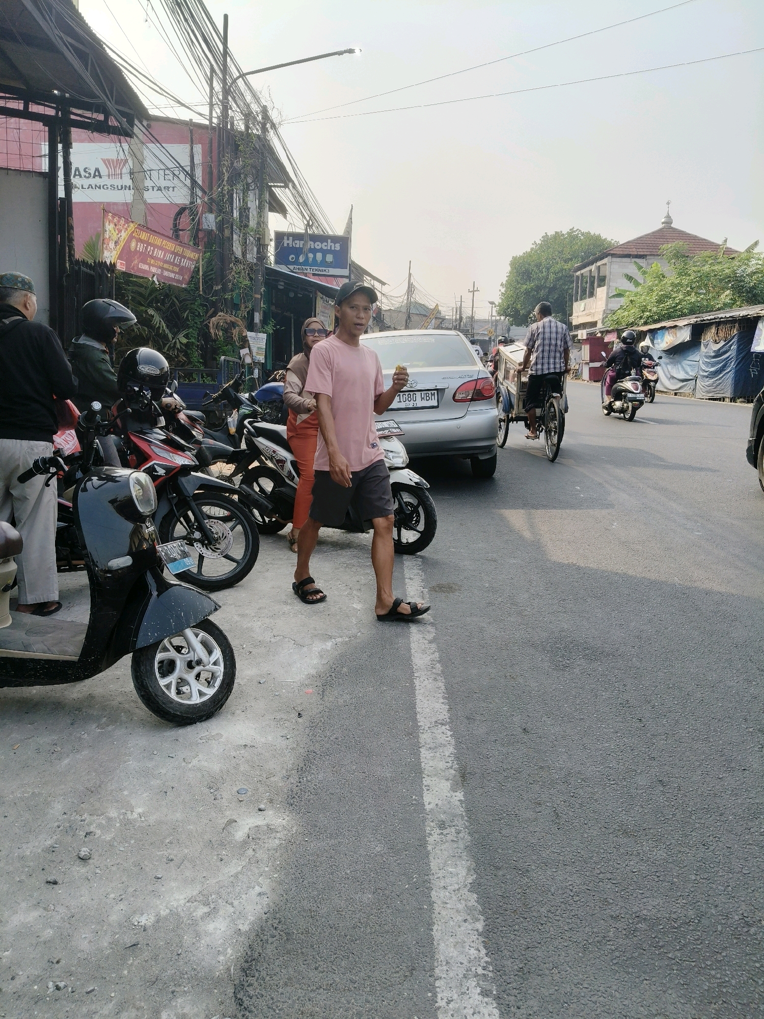In South Tangerang, Indonesia, a street scene unfolds with a mix of parked motorcycles and ongoing traffic. The foreground features a black scooter and several other motorcycles parked on a concrete surface next to a road. A young man in a pink t-shirt and dark shorts walks towards the viewer, holding a small item in his right hand. To his left, a woman in an orange outfit stands near a parked motorcycle. Behind them, a silver car with the license plate "1689 WBM" is stopped on the road. Further down the street, a man on a bicycle with a large cargo box is visible, along with several people riding motorcycles. Buildings line the left side of the street, displaying signs such as "ASA ENTERY" and "Hannochs." Overhead, a complex network of electrical wires crisscrosses the sky, and a street lamp is present. The sky is bright and overcast, suggesting daylight. The overall environment appears to be a busy urban area with a mix of commercial establishments and residential structures.