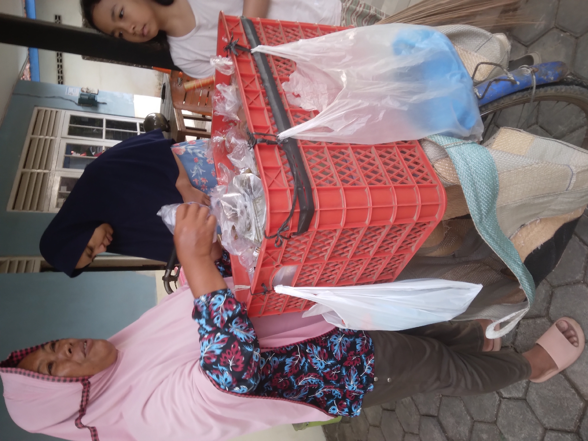 The image depicts a street scene in Desa Pamijen, Kecamatan Sokaraja, Indonesia. In the foreground, an adult woman wearing a pink hijab and a patterned shirt with blue and red floral motifs is interacting with a red plastic crate. The crate is filled with various items wrapped in plastic. Next to her, partially visible, is another woman in a dark hijab. To the right, a young girl in a white t-shirt is looking away from the camera. The crate is attached to a bicycle, which also has several bags, including white plastic bags and a woven bag, hanging from it. A broom is leaning against the bicycle. The ground is paved with hexagonal tiles. The background shows a building with white-framed windows and doors. The overall setting suggests a commercial transaction or distribution taking place outdoors.