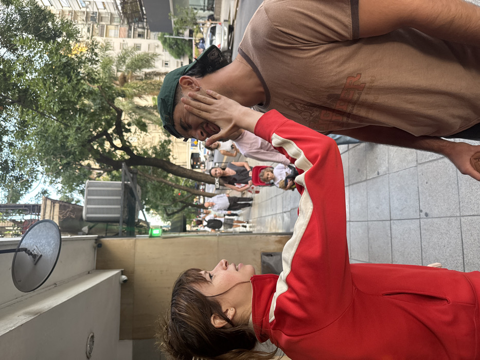 The image captures an urban sidewalk scene in Buenos Aires, Argentina, during daylight. In the immediate foreground, a light-skinned female with brown hair, wearing a red track jacket with white shoulder stripes, has her left hand pressed against the right cheek of a light-skinned male. The male, wearing a backward dark green baseball cap and a brown t-shirt, exhibits a contorted facial expression resulting from the pressure. His right hand holds a brown cylindrical object.
In the midground, several pedestrians walk past on the light-colored tiled sidewalk. Notably, a woman pushes a red stroller containing a small child with curly light-colored hair. The background features multi-story urban buildings consistent with Buenos Aires architecture, including one with an ornate balcony and an external air conditioning unit. Trees with green foliage line the street, and vehicles are visible further down. A convex security mirror is mounted to the side of a building above the foreground subjects, alongside a small green rectangular sign.