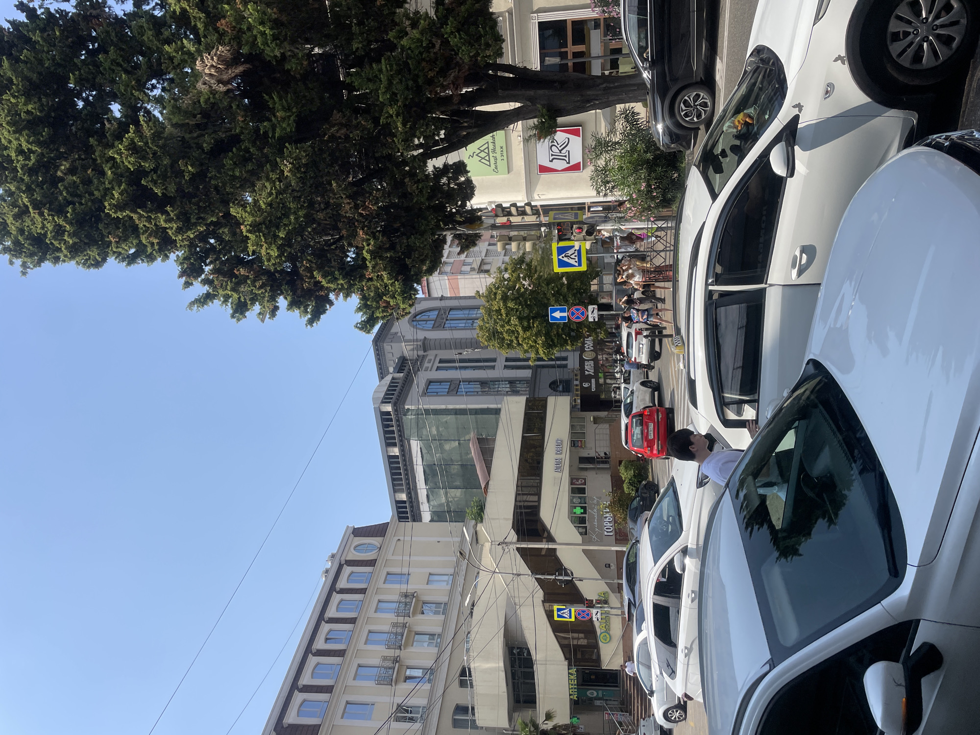 A sunny day in Sochi, Russia, shows a street scene dominated by several cars and buildings. In the foreground, a white car is parked, its windshield reflecting the surroundings. To its right, another white car is occupied by a person. Several other vehicles, including a red car and a black SUV, are visible on the street.
The background features a multi-story building with balconies and a modern glass-fronted structure. A large, mature tree stands tall on the right side of the image, its branches reaching towards the clear blue sky. Traffic lights and street signs are present, indicating an urban setting with regulated traffic flow. A group of people are seen walking on the sidewalk, some are waiting to cross the street. The overall atmosphere suggests a bustling city environment on a pleasant day.