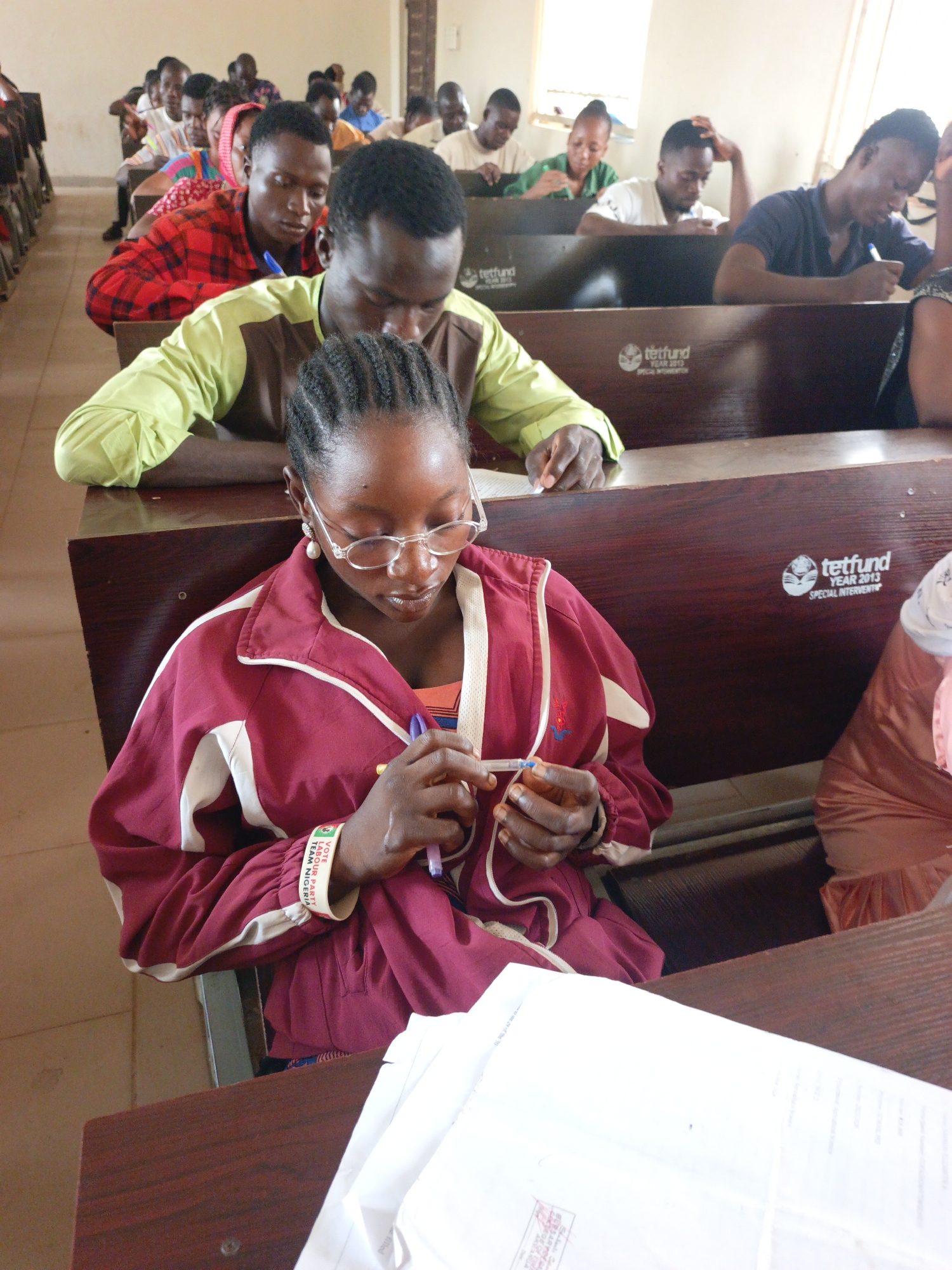 In a classroom setting in Akwanga, Nigeria, a group of students are seated at wooden desks, seemingly engaged in a task, possibly an exam or assignment.
The focus of the image is a young woman in the foreground, wearing glasses and a maroon jacket with white stripes. She is holding a pen and looking down at something in her hands, possibly fiddling with it. She is wearing a bracelet with the word "LABOUR PARTY" and the phrase "VOTE TEAM NIGERIA."
Behind her, a male student is wearing a green and brown shirt. Further back, additional students are seen in the classroom, some writing or reading.
The desks in the classroom have the "tetfund YEAR 2013 SPECIAL INTERVENTION" logo printed on them. There are white papers on the desks in the foreground, suggesting a test or assignment is in progress. The room is well-lit with light coming in from a window.