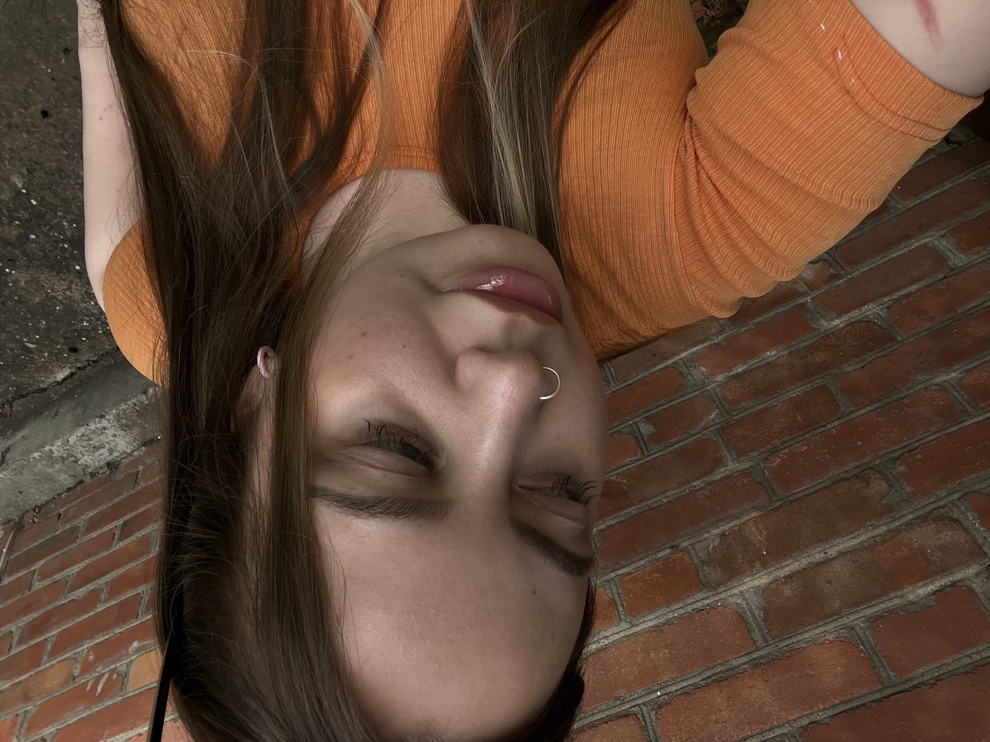 This is a portrait of a young person lying down, with their head tilted back. They are wearing an orange, ribbed top. Their long, brown and blonde hair falls around their face. They have a silver hoop piercing in their nose and a pink stud earring. Their eyes are closed, and their lips are slightly parted. The background consists of a red brick wall and a darker, rough surface that appears to be concrete or pavement. The lighting suggests it could be daytime, possibly outdoors, given the brick and concrete elements. The overall mood is relaxed and perhaps contemplative. There is no visible text in the image.