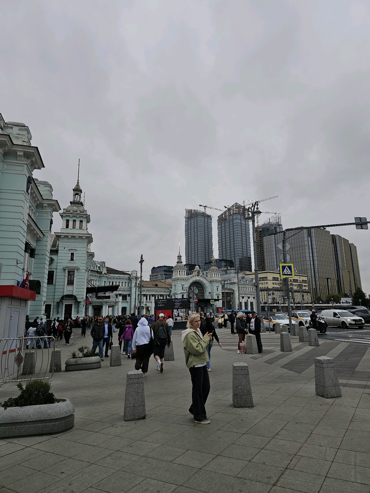 The image depicts a street scene in Moscow, Russia, likely in front of Komsomolskaya Square, also known as "Three Station Square," due to the prominent architecture and the presence of train stations.
In the foreground, a woman is standing and looking at her phone. She is wearing a light green jacket and dark pants. To her left, several concrete planters with sparse vegetation are arranged along the pedestrian walkway. The walkway is paved with large square tiles.
A crowd of people are walking in various directions across the pedestrian area. They are dressed in casual clothing.
In the midground, a grand, light blue building with ornate architectural details and a prominent clock tower stands on the left. This building appears to be a historic train station. Behind it and further to the right, modern skyscrapers are under construction, with cranes visible against the overcast sky. Another, more contemporary building with a glass facade is situated on the far right.
The street to the right of the pedestrian area is busy with traffic, including cars and a motorcycle. Traffic lights and road signs are visible. The sky is cloudy and gray, suggesting an overcast day. The overall atmosphere is that of a bustling urban environment with a blend of historical and modern architecture.