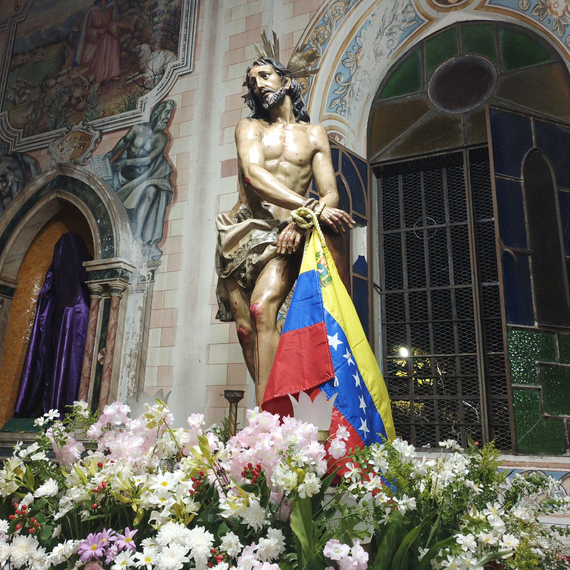 The image is taken inside a church in Tinaquillo, Venezuela. In the foreground is a statue of Jesus Christ with hands bound, draped with the Venezuelan flag. A large arrangement of flowers surrounds the base of the statue. To the left is a niche with a purple draped figure. A mural adorns the wall in the background, and a stained-glass window is to the right.