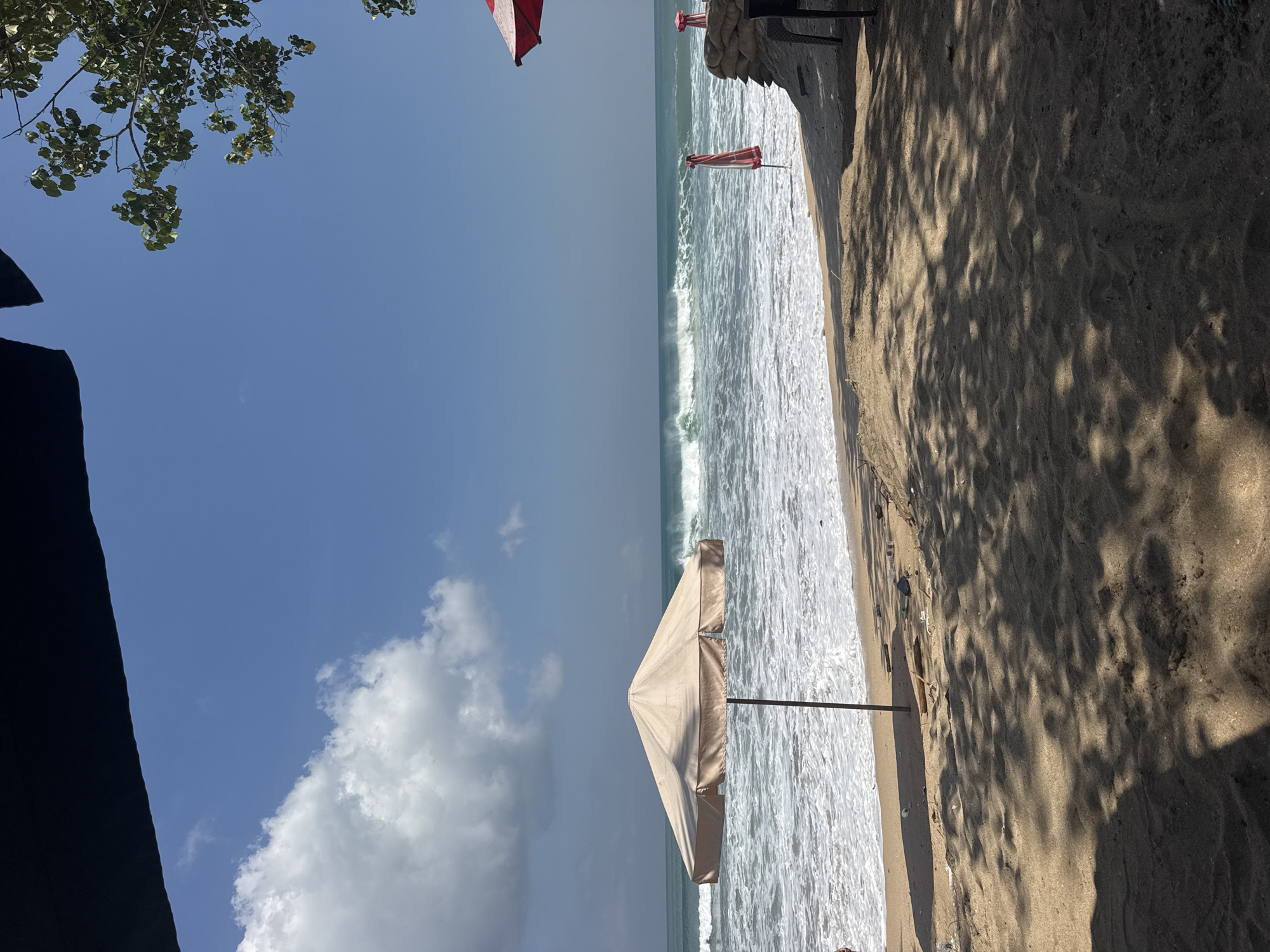 A beach in Kuta, Indonesia, faces the ocean under a clear blue sky with scattered clouds. Foreground details include sandy ground with dappled shade from unseen trees, and a beige beach umbrella planted in the sand. The middle ground shows the ocean with moderate waves breaking and white foam washing ashore. Two red and white striped beach umbrellas are visible near the water's edge. In the background, the ocean stretches to the horizon, meeting the sky. The overall scene depicts a relaxed, sunny day at the beach.