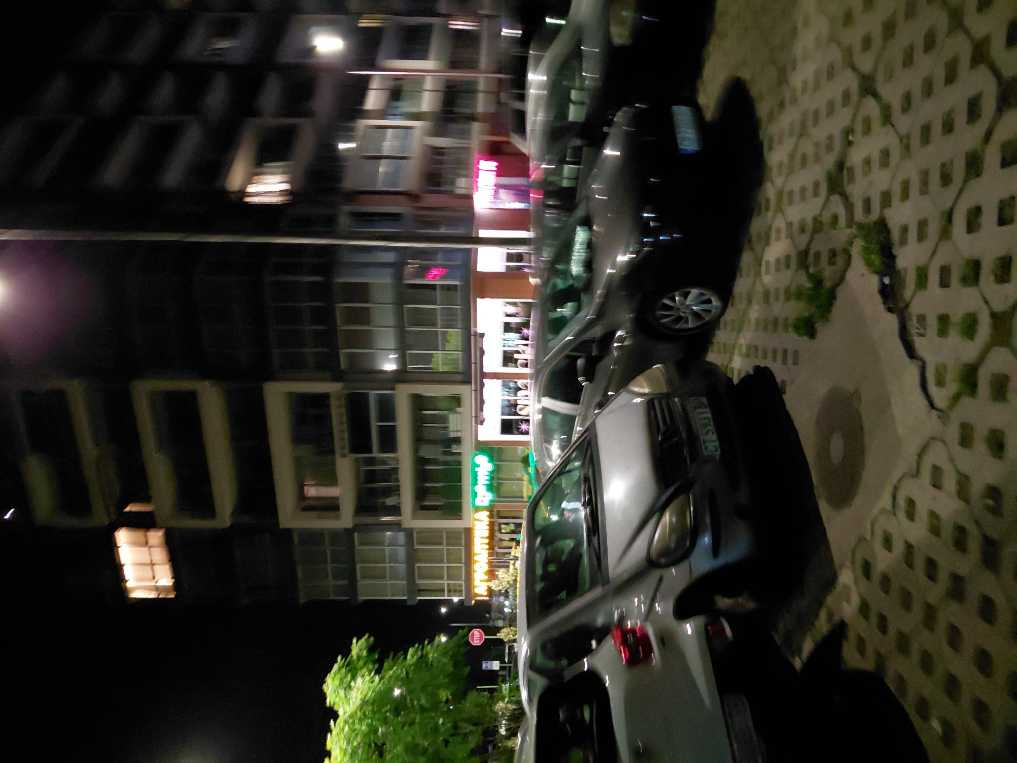 An outdoor, nighttime scene depicts a row of parked cars in front of a modern, multi-story building. The building features a facade with large glass windows and illuminated sections. A neon sign in green letters, reading "АГРОАПТЕКА," is visible on the building's ground floor, alongside a pink neon sign. One of the parked cars, a silver Mercedes-Benz A-Class, is in the foreground and has the license plate "CT 5931 B". Adjacent to the cars is a parking lot paved with decorative concrete blocks, some of which have grass growing through them. A street lamp illuminates the scene, casting shadows from the cars. A red "STOP" sign is visible in the background, near a tree with green foliage. The overall atmosphere is urban and commercial.