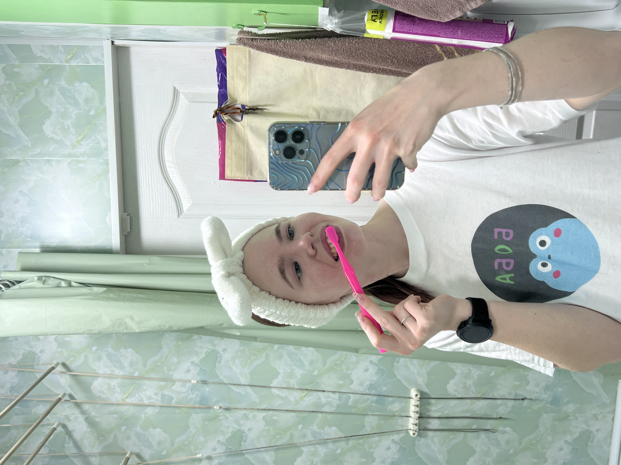 A young woman with a white rabbit ear headband is taking a selfie in a bathroom, brushing her teeth with a pink toothbrush. She is holding a smartphone with a blue patterned case. She's wearing a white t-shirt with a cartoon character on it. On her left wrist, she's wearing a black smartwatch. Her nails have a light pink design. The background includes a tiled bathroom wall, a white cabinet, and a retractable clothesline. Based on previous information, this scene is located in the city of Chaikovsky, Russia.