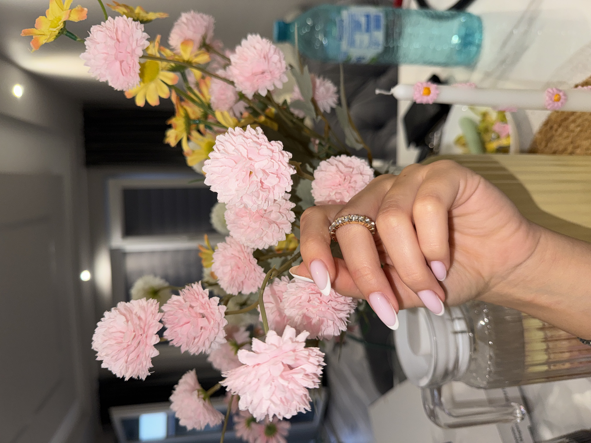 A person's hand with a gold ring and French manicured nails is positioned amidst a bouquet of pink and yellow flowers. A water bottle and a decorated candle are visible in the background, resting on what appears to be a wooden surface. The location is Burgas, Bulgaria.