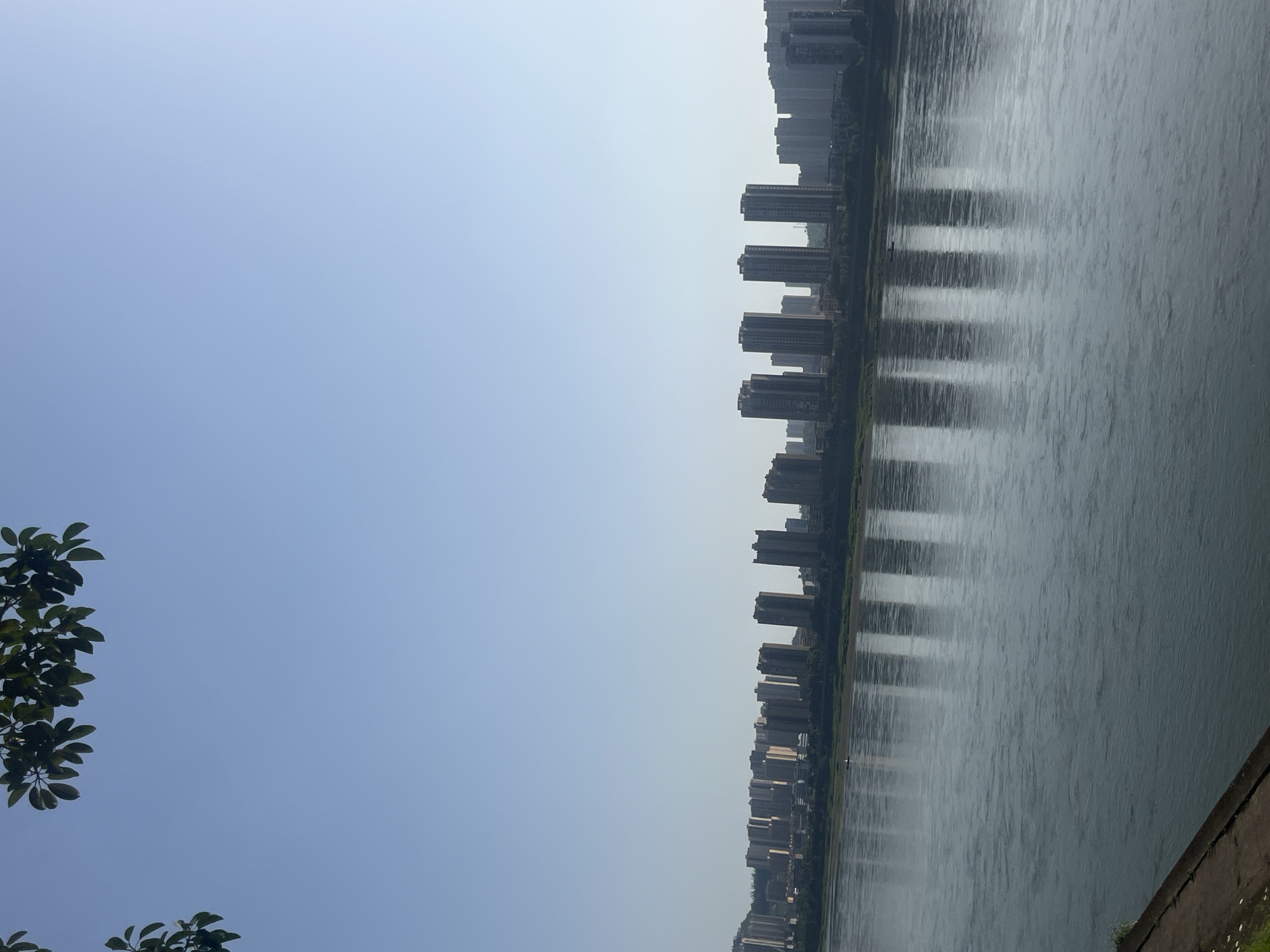 The image, taken in Jiangjin, China, presents a wide panoramic view of an urban river scene on a clear day. The frame is horizontally divided into two main sections: a vast, clear sky on the left and a river with a city skyline on the right.
The left half of the image is dominated by a bright, light blue sky, which transitions to a slightly paler, almost white hue towards the horizon line where it meets the distant city. There's a hint of haze in the distance, but the overall impression is one of brightness and openness. In the bottom-left corner, dark green foliage, consisting of several leaves and branches, partially frames the shot, adding a natural element to the foreground.
The right half of the image features a wide body of water, likely a river, reflecting the sky and the urban landscape. Along the far bank of the river, a dense and continuous line of tall, modern high-rise buildings forms a distinct city skyline. These buildings are predominantly rectangular and appear residential or commercial, stretching far into the background. Their reflections are clearly visible on the water's surface as elongated, blurred vertical strips, suggesting calm water with slight ripples. The immediate riverbank in the lower right is a dark, solid structure, possibly a concrete embankment, with minimal vegetation.
No people or specific activities are discernible, giving the scene a quiet and contemplative atmosphere. The time of day appears to be daytime, with natural, soft lighting indicating possibly late morning or early afternoon. There is no visible text in the image.