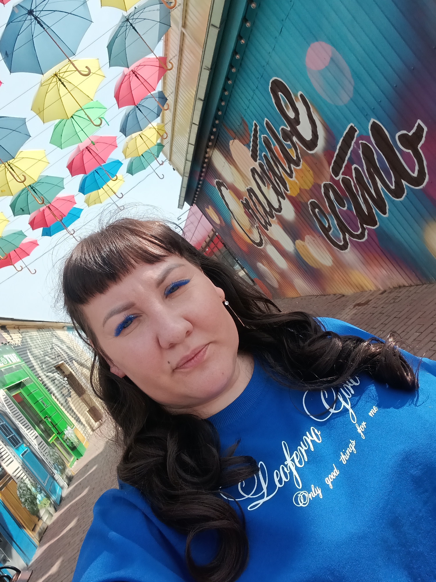 A woman with dark hair and blue eyeliner is the main subject of a selfie taken in Osa, Russia. She is wearing a bright blue sweatshirt with white cursive writing. Above her head are rows of colorful umbrellas suspended in the air. To her right is a blue wall with graffiti. To her left are buildings with brightly colored doors. The ground is paved. The sky is bright and clear.