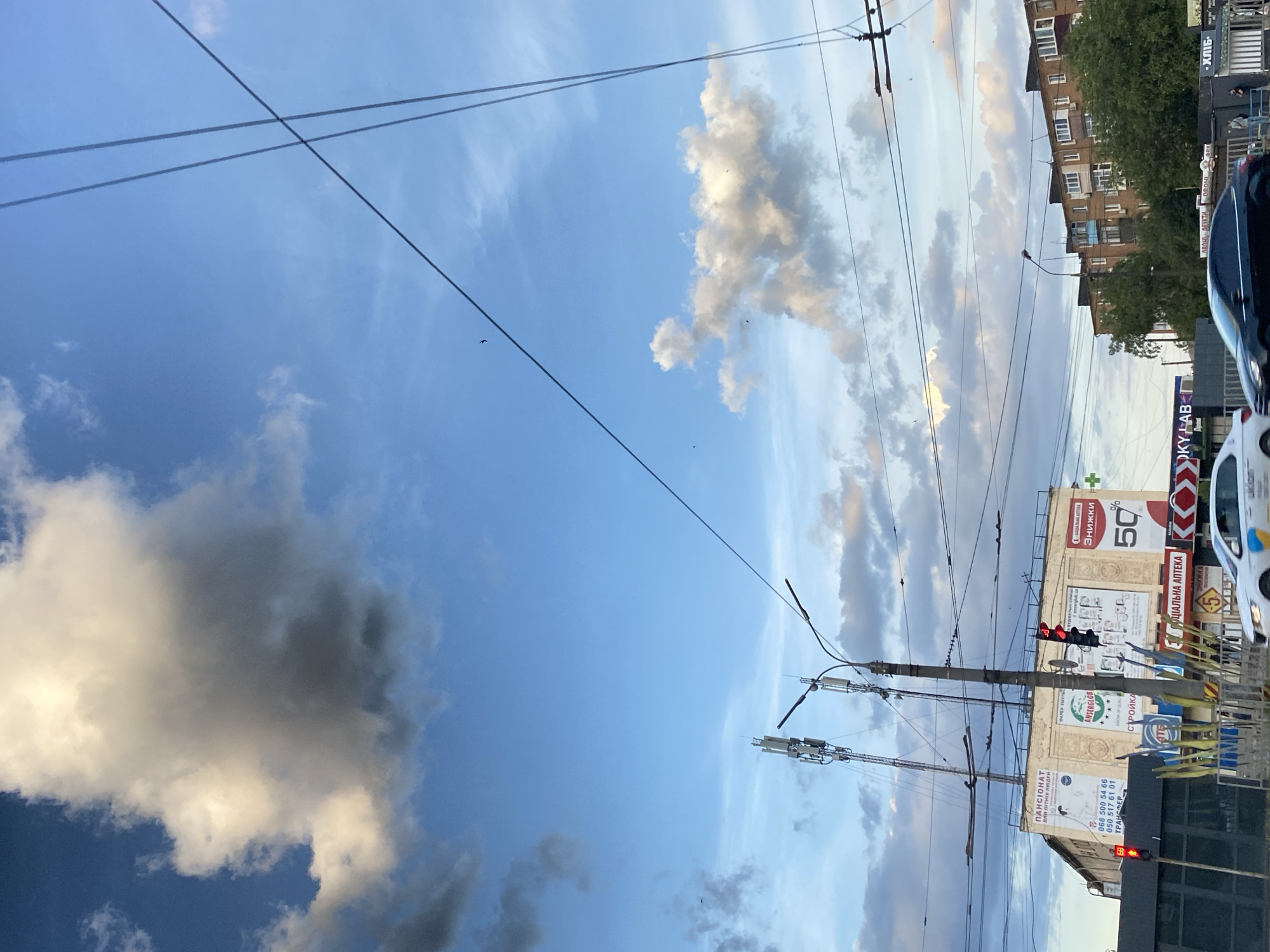 In an urban setting under a blue sky with scattered clouds, an intersection is visible. A traffic light displays red. Several utility poles and communication towers are present, along with numerous overhead wires. Buildings line the street, including one with advertisements for a "PANCIANAT" and "SOCIAL APTEKA" offering "ZNISHKY 50%". The foreground shows a street with cars, including a white car with Ukrainian flag markings and a black sedan, in motion. Trees and a low barrier are also visible. Small birds can be seen flying in the sky.