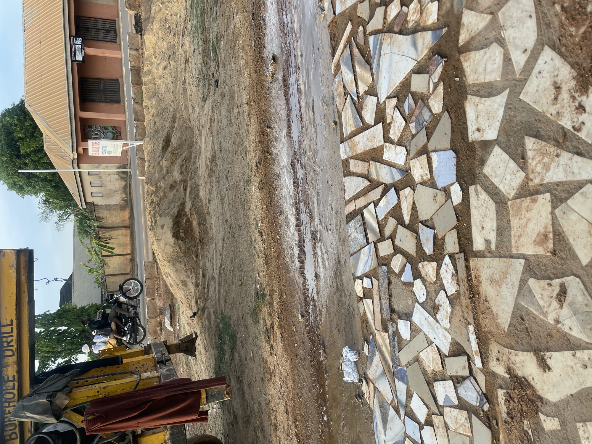 The image displays an outdoor scene, likely a street or unpaved area in Narayi, Nigeria, captured during the daytime under a clear sky. The perspective is tilted, with the ground surface dominating much of the frame.
In the foreground, a large yellow "BOREHOLE DRILL" machine is prominent, with red trousers and a grey garment draped over its components. The ground itself is a mix of sandy dirt and an informal path or decorative element created from numerous irregularly shaped, light-colored broken tiles or ceramics embedded in the earth. A muddy, wet track or channel runs through the sandy portion of the ground.
Several individuals are visible in the midground, particularly around a black motorcycle with white wheels. Two people wearing white shirts are discernible near the motorcycle.
In the background, there are several buildings and sparse vegetation. A reddish-brown building with a corrugated metal roof features barred windows. Below these windows, graffiti with the letter "R" is visible on the wall. In front of this building, a white rectangular sign on a pole reads "ALAN PANTIES COSMETICS" and "WE SELL ALL KIND OF HAIR WEAVES WIGS" followed by a phone number starting with "0803". A smaller, dark sign is also affixed to the building's wall. Behind the main building, another lighter-colored structure and green trees are present.
The overall scene suggests a functional, possibly commercial or utility-focused environment, with the borehole drill indicating construction or water access work.