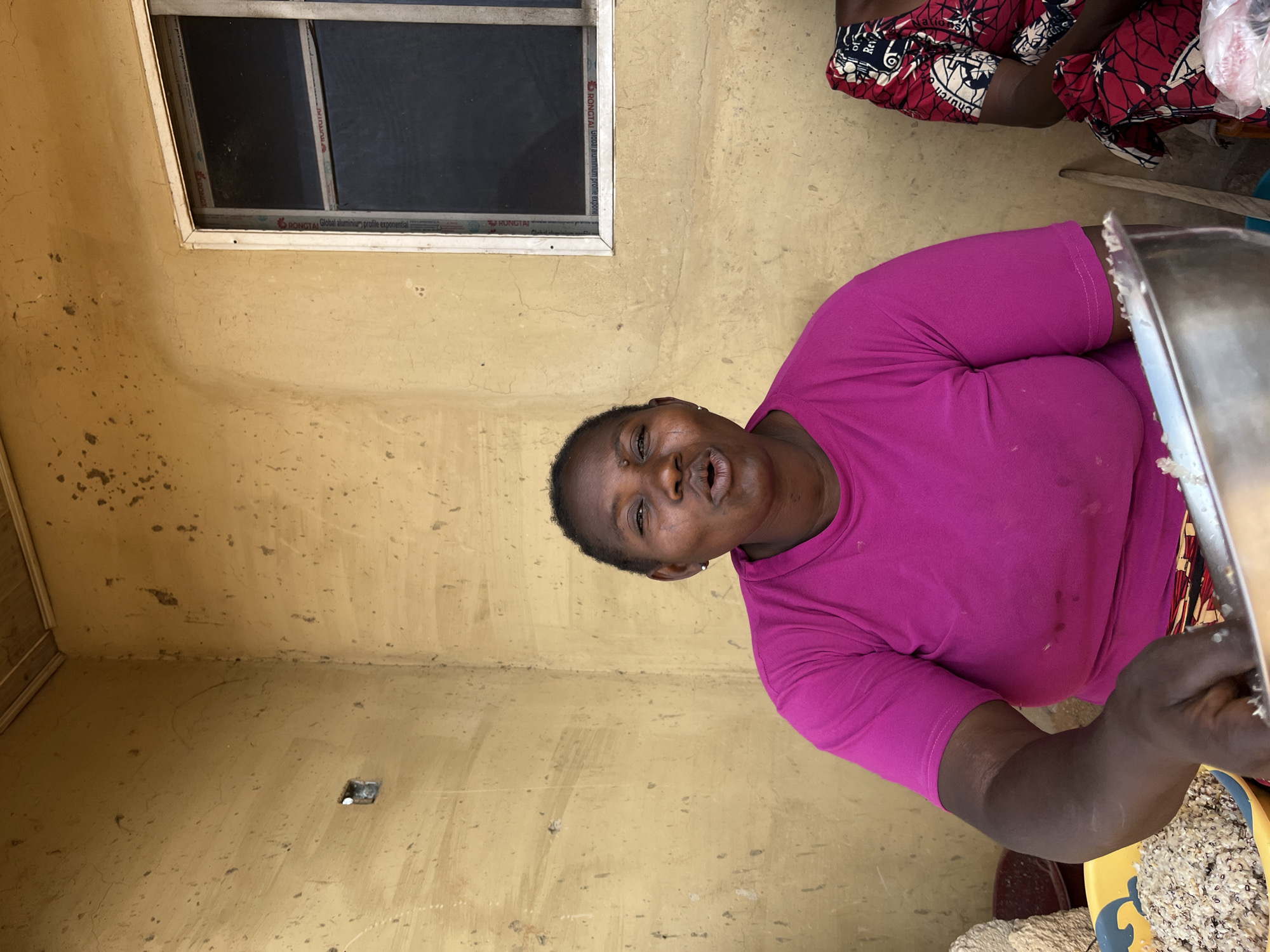 In the city of Jos, Nigeria, a woman is captured in a close-up, seated within a rudimentary indoor or semi-outdoor space. The dominant subject, a woman of African descent, occupies the central and lower-left portions of the frame, wearing a solid, bright magenta short-sleeved t-shirt. Her expression is animated, with her mouth slightly open, suggesting she is speaking or reacting. A small, light-colored stud earring is visible in her left ear. Her dark, short hair is pulled back or closely cropped.
Her left arm and hand are visible in the lower-left foreground, actively engaged with food preparation. Her hand is positioned near a large, silver-colored metal bowl. To her left, a yellow plastic bowl, featuring a blue dolphin or fish design, is filled with a mixture of light-colored grains, possibly rice and beans.
To the right of the primary subject, a second person is partially visible, only their right shoulder and arm in frame. This individual is wearing a red and white patterned fabric, possibly an ankara print, with the text "Church of Rev" partially discernible on the fabric. A thin wooden pole leans against the wall between the two individuals.
The background consists of a textured, matte yellowish-brown wall, showing various cracks, darker stains, and splatters, particularly prominent in the upper corners. On the upper right, a rectangular window with a white frame is set into the wall. The window panes are dark, possibly screened, and feature internal mullions. Legible text on the window frame includes "RONGTAI" and "aluminum profile." In the upper-left portion of the wall, a small, square, dark recess or hole is visible. Another lighter-colored, fibrous object, possibly a sack, is partially seen on the far left, behind the yellow bowl. The scene is evenly lit, indicating natural daylight.