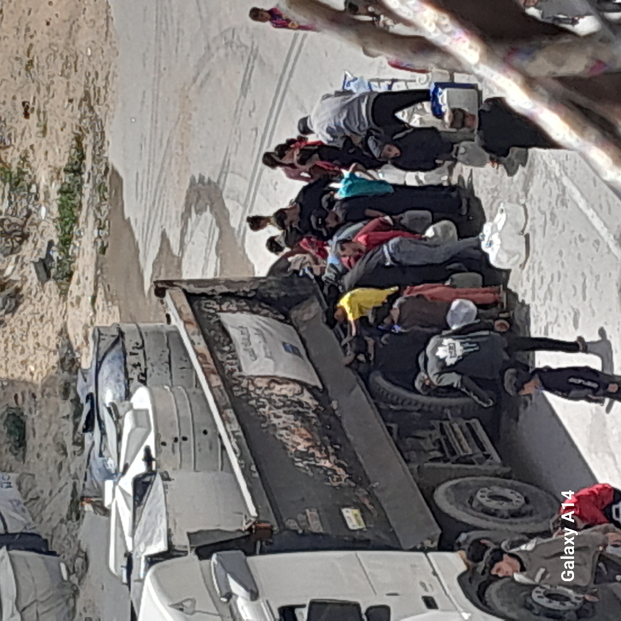 The image shows a group of people lying down on and around the back of a large, white truck in what appears to be an urban or semi-urban area. The ground is a mix of pavement and dirt. Some people are lying on the flatbed of the truck, while others are on the ground beside or underneath it. Clothing colors vary, with some individuals wearing yellow, red, gray, and other assorted colors. The truck seems to have a damaged or worn flatbed. There is also trash and debris visible in the background on the dirt area.
