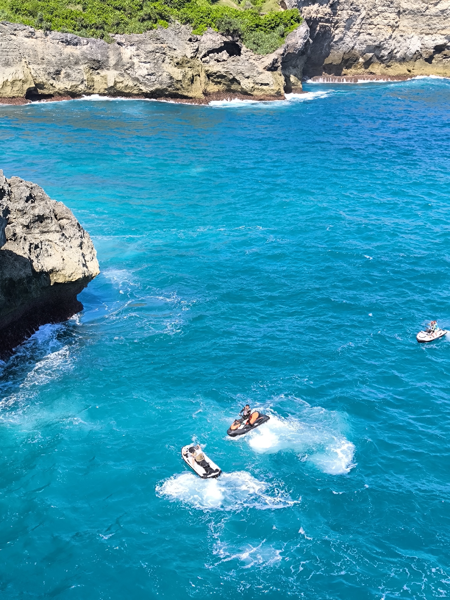 The image captures a scene at Nusa Penida, Indonesia, featuring the turquoise waters of the Indian Ocean and rugged coastal cliffs. The top of the frame shows a section of the cliff face covered in green vegetation. The focus is on the water, where three jet skis are in motion. Two of the jet skis are closer to the viewer, each with one person riding them. The third jet ski is farther away and appears to have two occupants. White wakes indicate the path of the jet skis across the blue water. In the bottom-left corner, a rocky outcrop adds to the ruggedness of the landscape.