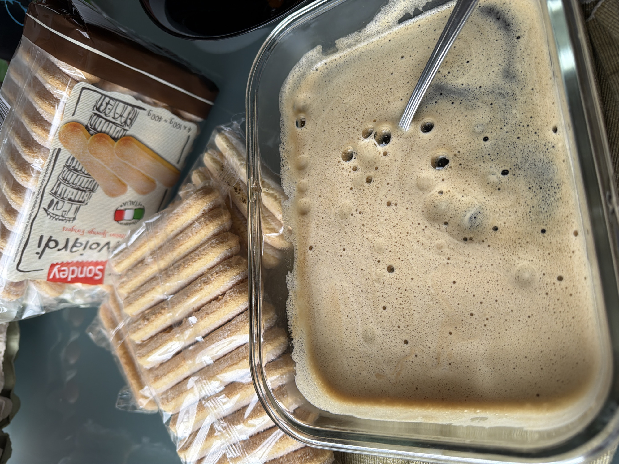 The image shows ingredients for making tiramisu. In the foreground is a rectangular glass dish filled with a brown, foamy liquid, presumably coffee. A metal spoon rests inside the dish. To the left, there are two unopened packages of Sondley Voiardi ladyfingers, with the Italian flag visible on the packaging. The setting appears to be indoors, possibly a kitchen. There is no clear evidence indicating the location is specifically Sofia, Bulgaria.