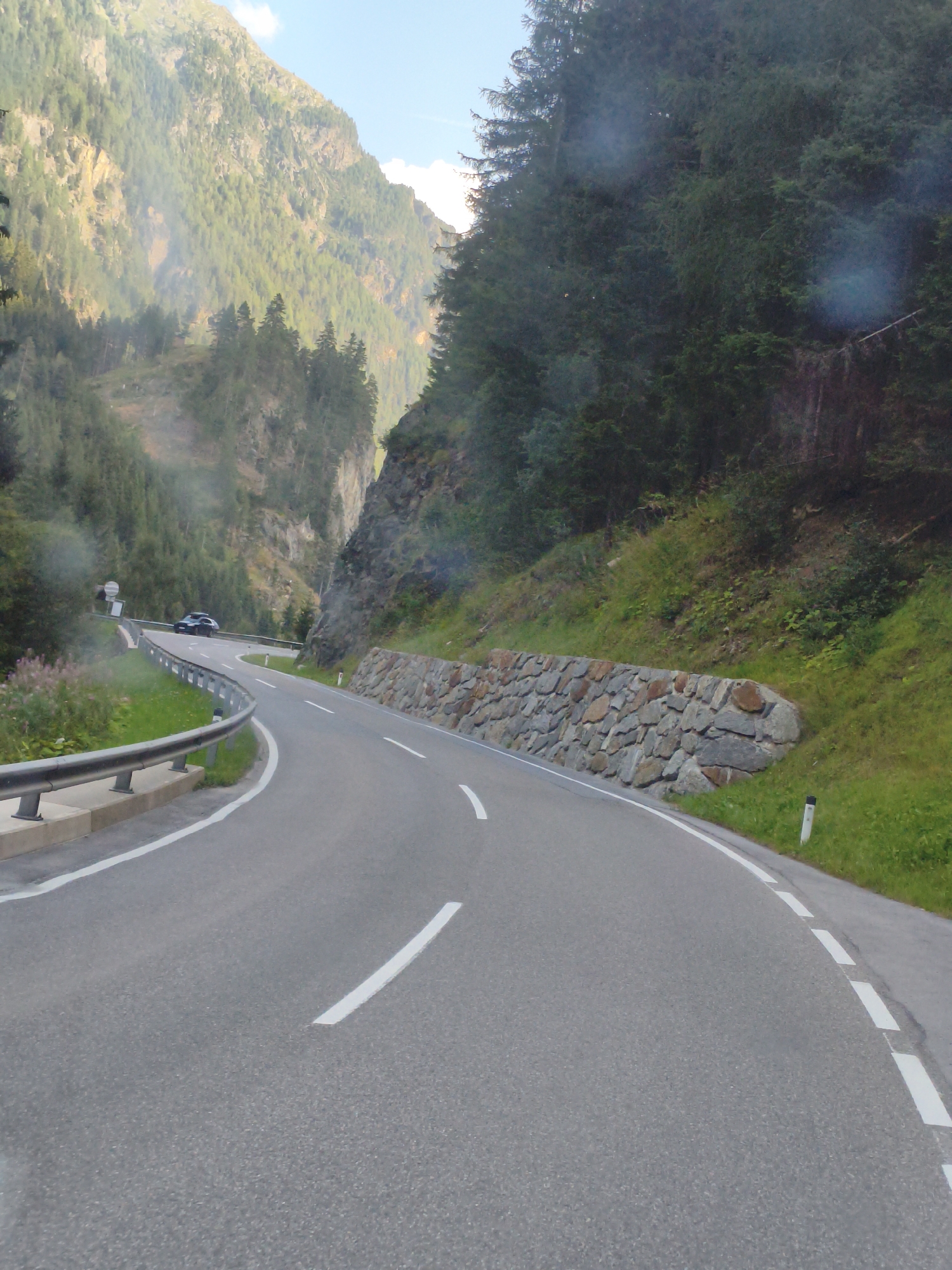The image shows a winding asphalt road passing through a mountainous, forested area in Sölden, Austria. The road has white lane markings, and a metal guardrail runs along the left side. On the right side of the road, a stone wall has been constructed to retain the hillside. Vegetation, including trees and grass, covers the slopes. The sky is visible in the upper part of the image. A dark-colored car is seen driving on the road in the distance.
