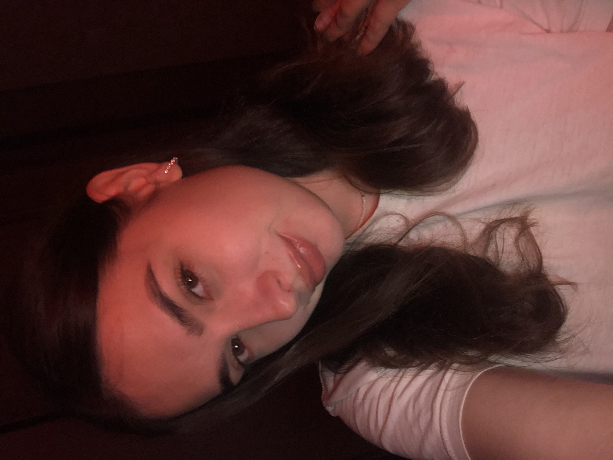 A young woman with long, dark brown hair and brown eyes is looking at the camera, her head tilted to the left. She is wearing a white t-shirt and a small, sparkly earring in her left ear. The lighting is dim and reddish, casting a warm glow on her face. Her right hand is resting on her shoulder, and a dark background is visible behind her. The image appears to be a selfie taken indoors, possibly in a room or a dimly lit space. There are no indications of the location being in Noviy, Russia.