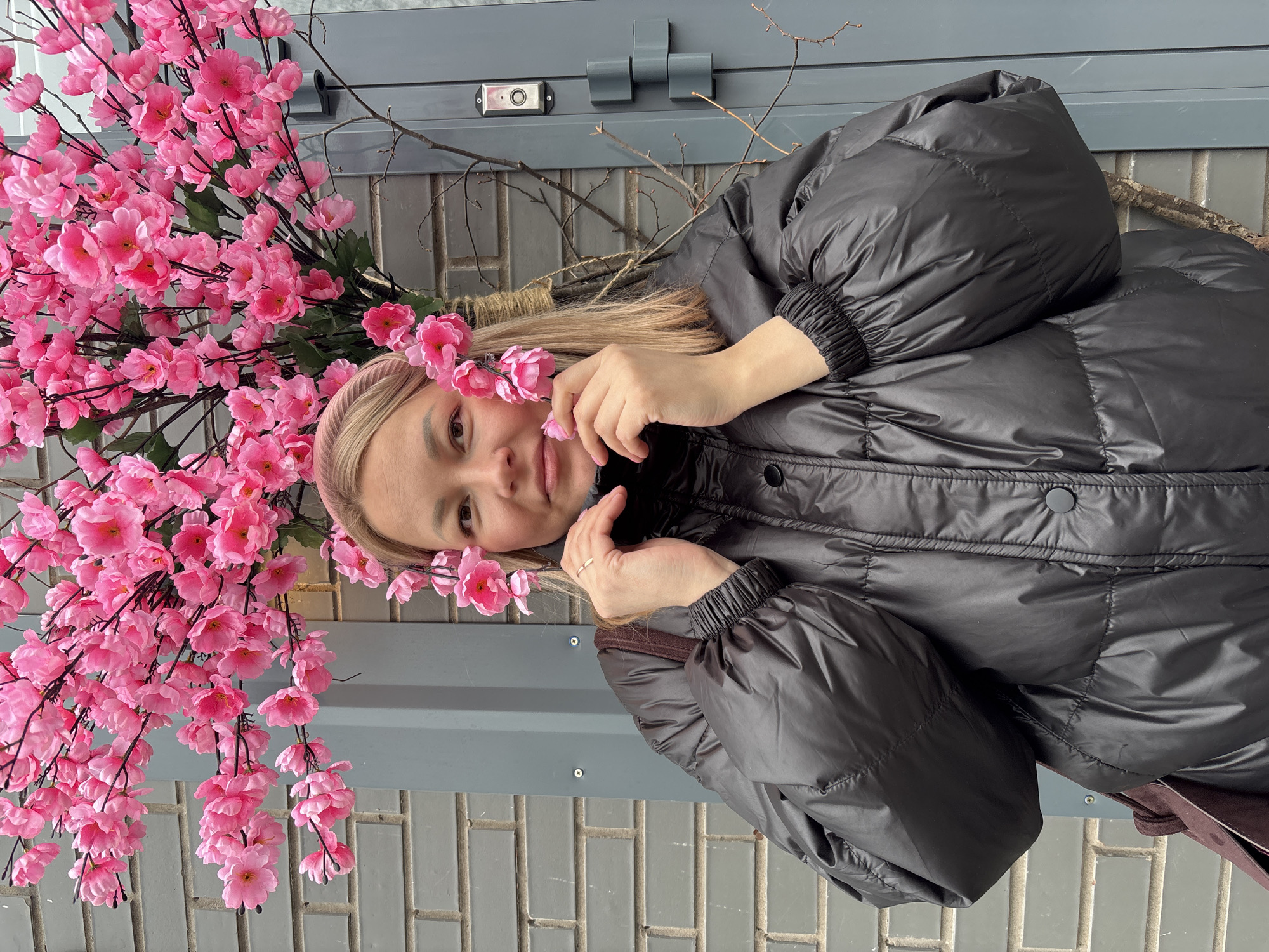 The image shows a woman in a black puffer jacket lying next to a wall decorated with pink artificial cherry blossoms. The wall appears to be made of grey bricks and is located outside. The woman is wearing a pink headband. The photo appears to have been taken in Izhevsk, Russia.