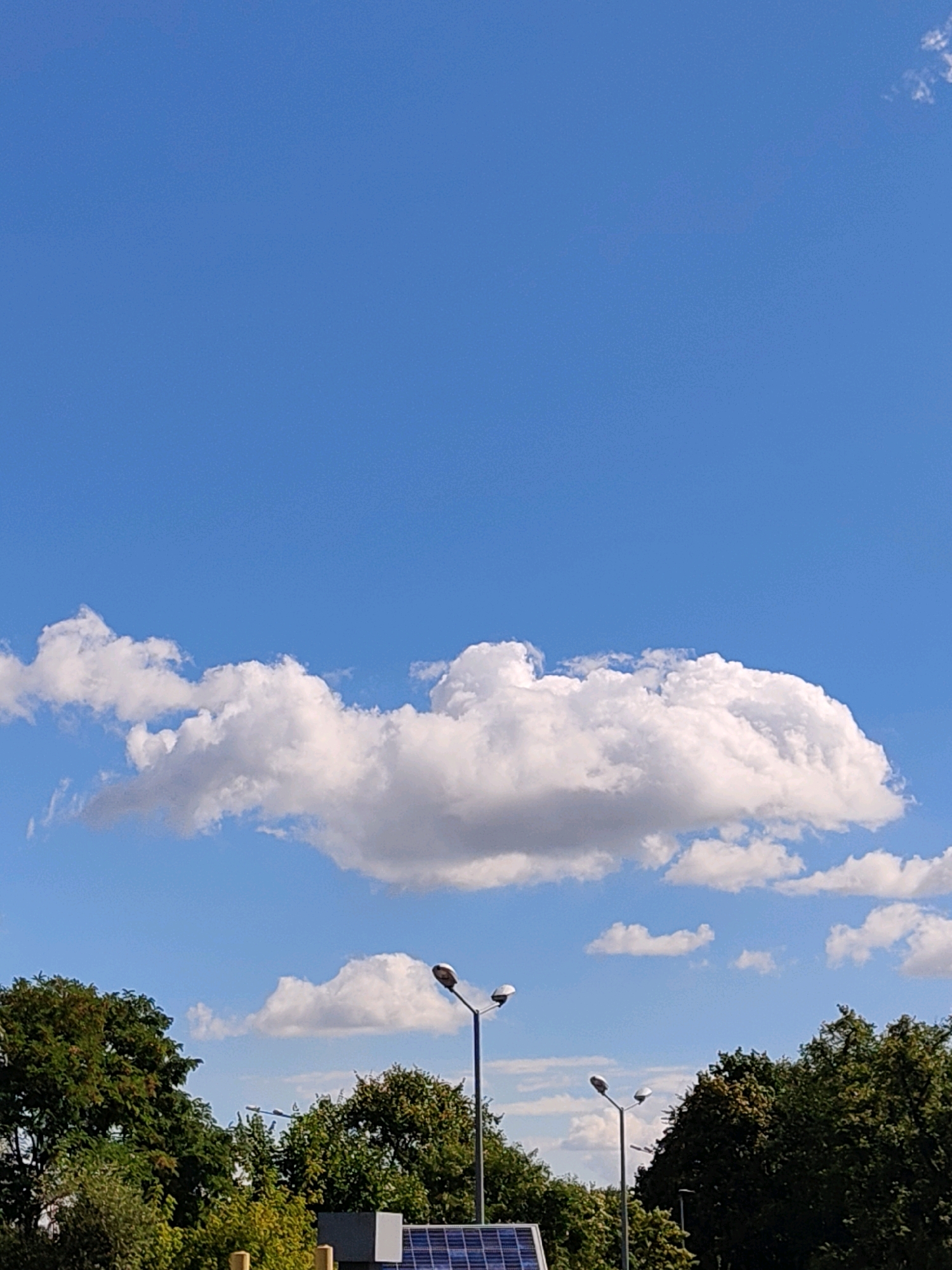 This image depicts a clear blue sky with scattered white cumulus clouds. The lower portion of the image shows a line of lush green trees, suggesting an outdoor environment. Two streetlights with modern designs are visible, one on the right and another further to the left, partially obscured by trees. In the foreground, a solar panel array is partially visible, indicating a sustainable energy installation. The overall scene suggests a bright, sunny day in an urban or suburban setting. While the image does not explicitly show any landmarks of Krakow, Poland, the presence of modern streetlights and solar panels could be indicative of public spaces or infrastructure developments in the city.