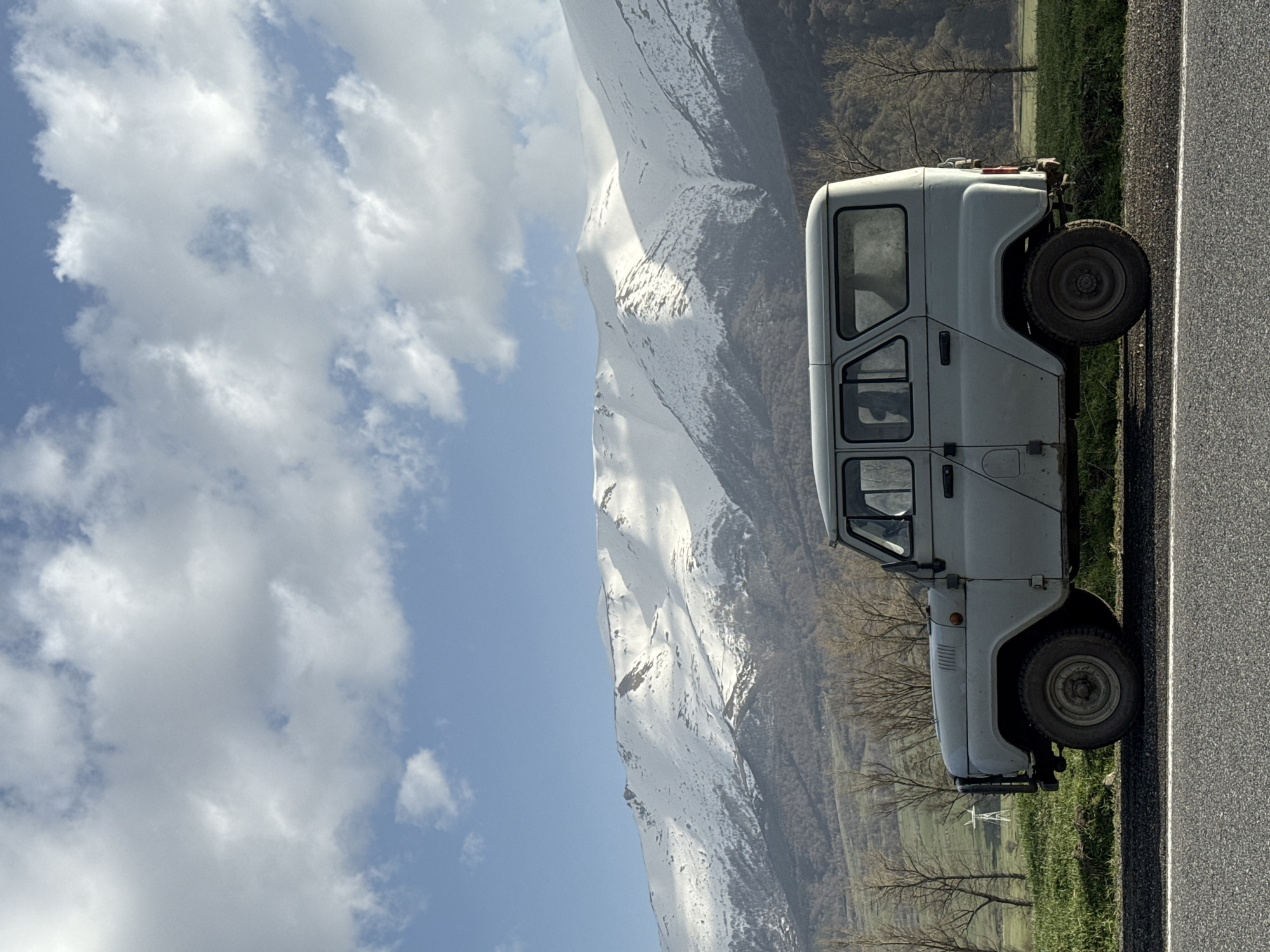 A white UAZ-452 van sits parked on the shoulder of a gray asphalt road in Margahovit, Armenia. The road runs parallel to a green grassy area interspersed with bare trees. Behind the van, a large mountain range, partially covered in snow, dominates the landscape, with a clear blue sky and puffy white clouds above the mountain.