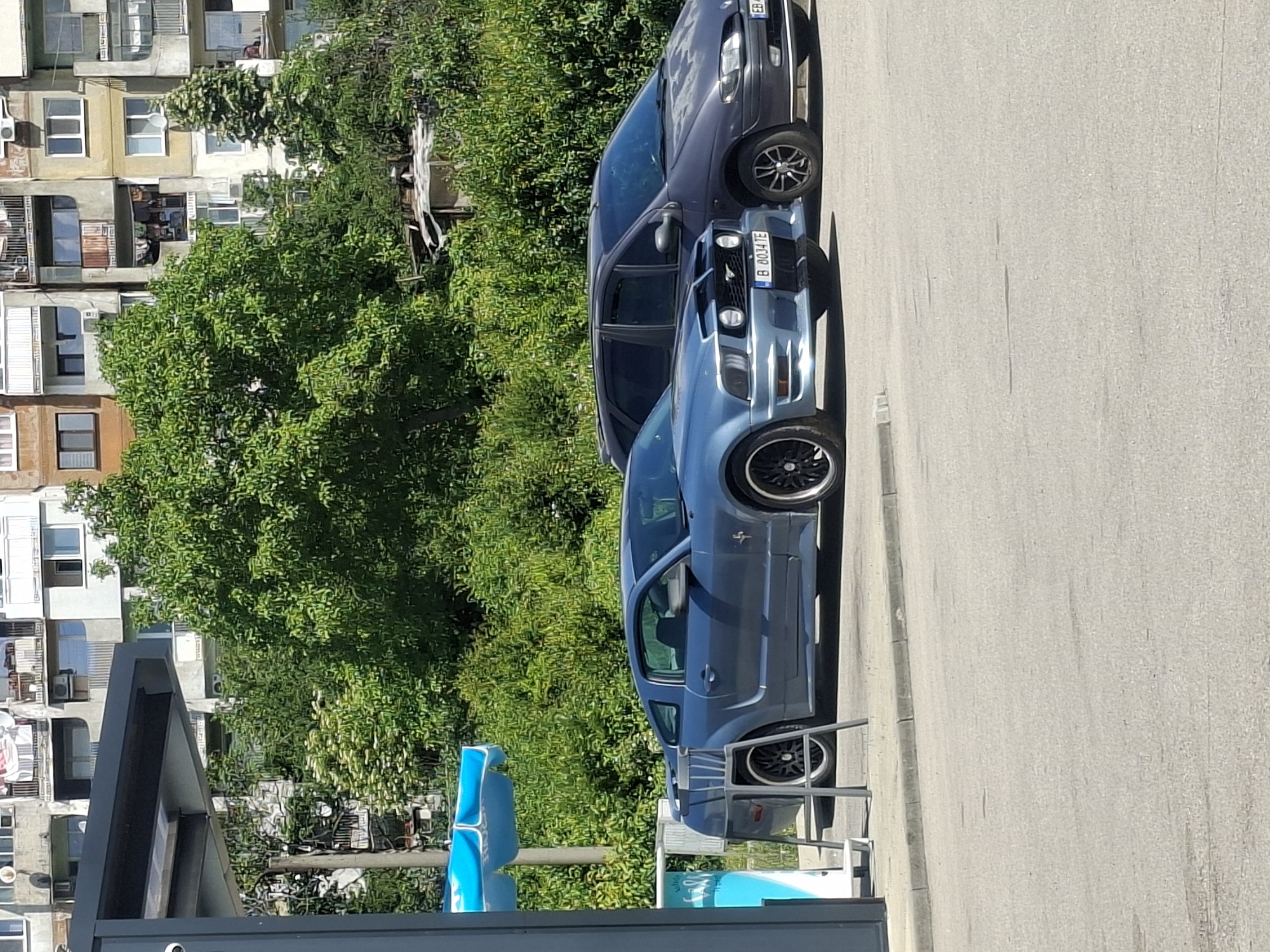 The image shows a street in an urban area, possibly a residential neighborhood in Unknown, Unknown. A blue Ford Mustang is parked on the right side of the street, behind a dark-colored sedan. Behind the cars, there is a large green tree and a multistory residential building. In the foreground, there is a partially visible structure with blue and white elements, potentially a bus stop or a shop front. The street is made of asphalt and shows signs of wear and tear. The sky is not visible in the picture. The license plate of the Mustang is partially visible and contains the characters "B 0004TE".
