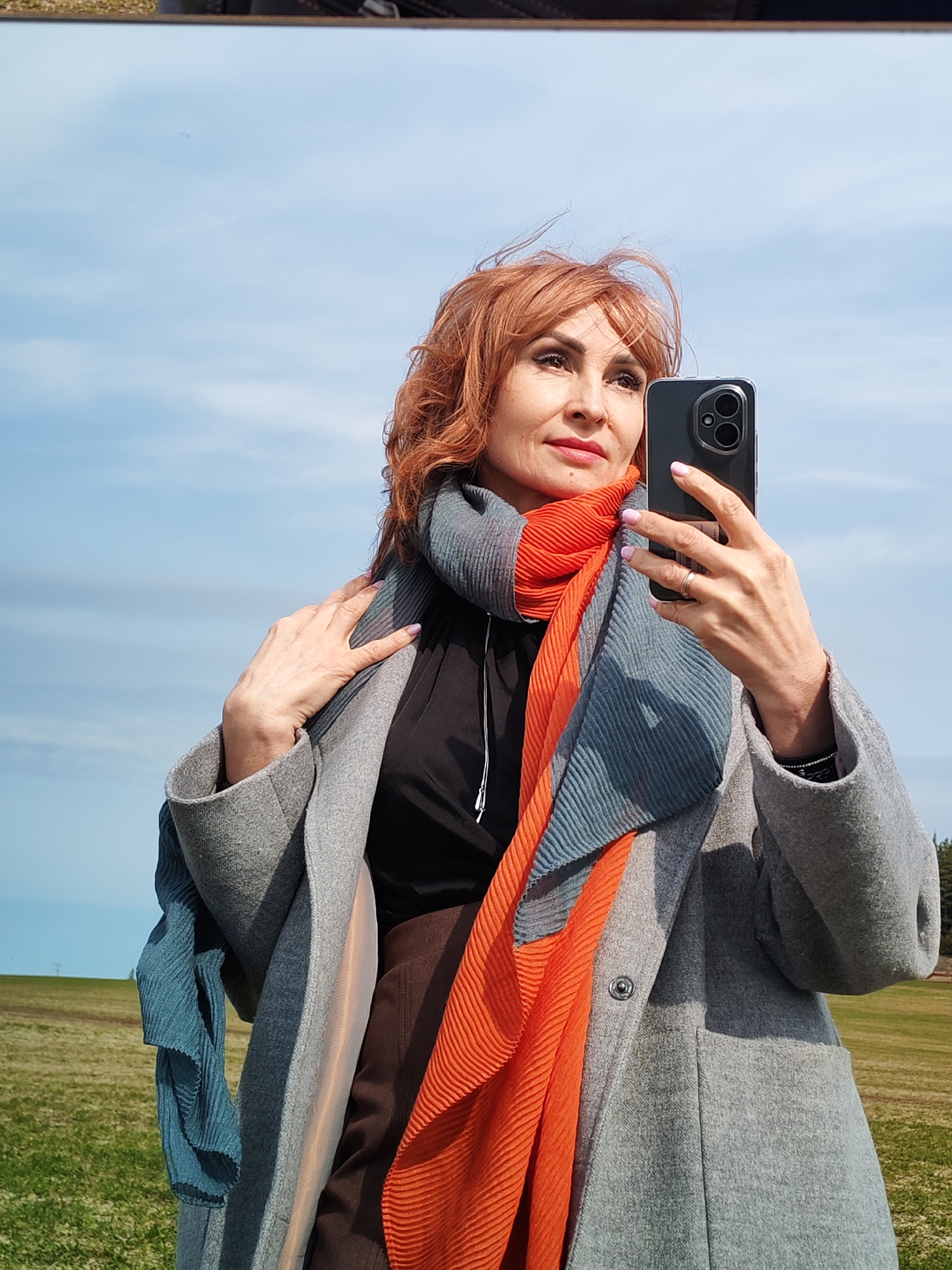 A woman with reddish-brown hair is holding a black smartphone, presumably taking a selfie outdoors. She is wearing a gray coat and a black shirt underneath, with an orange and blue pleated scarf wrapped around her neck. Her nails are painted a light pink color. She is standing in a field of golden grass under a blue sky with light clouds. The location is in Нечкино, Russia.
