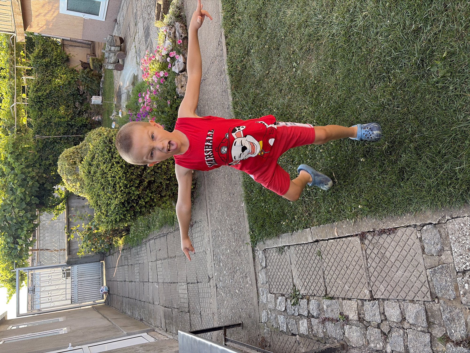In the city of Dolni Dabnik, Bulgaria, a young boy wearing a red Paw Patrol t-shirt and red shorts, along with blue Crocs, stands on a grassy lawn in a backyard. He is making a playful, exaggerated facial expression with his tongue sticking out and his mouth open. His arms are extended outwards, with his left arm pointing forward and his right arm bent at the elbow. The backyard features a paved walkway made of interlocking gray tiles and a section of cobblestones. Beyond the walkway, there are bushes, a wall, and a wooden cart. A metal table is partially visible on the left side of the image. The sun is shining, casting shadows and illuminating the scene with warm light.