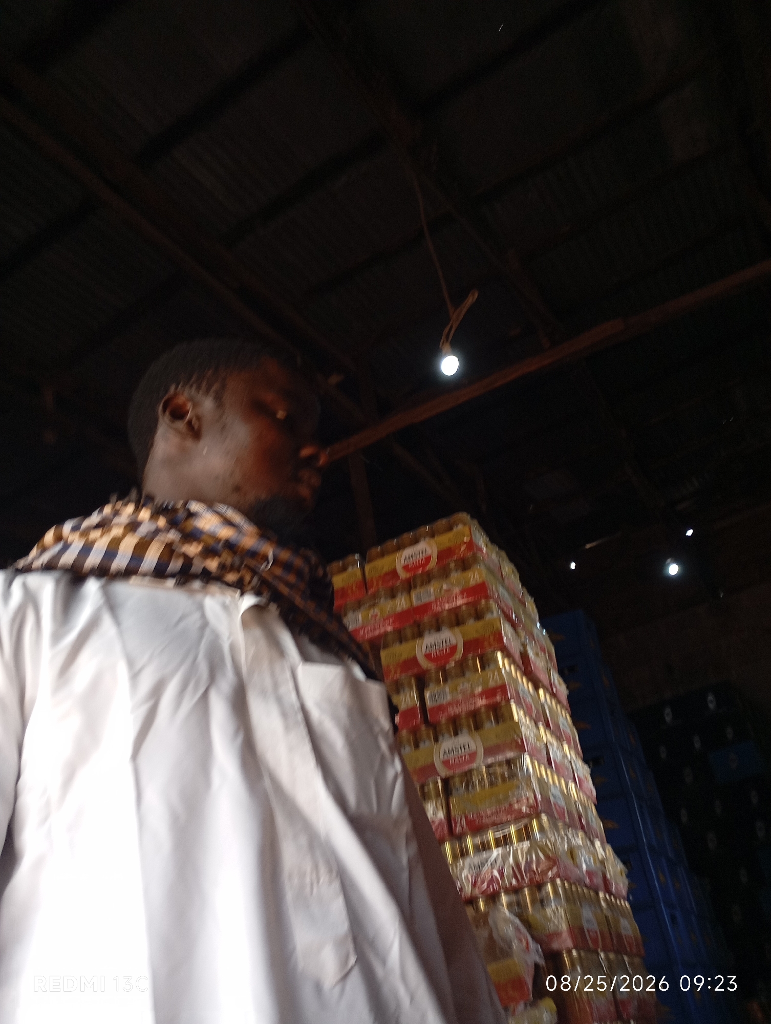 The image is a waist-up, close-up shot of a man in Shendam, Nigeria, wearing a white garment and a patterned scarf. He is looking away from the camera. Behind him, stacks of beer bottles in yellow and red packaging are visible, along with blue crates. The scene appears to be indoors, possibly a storage area or market, with a corrugated metal roof and visible wooden beams overhead. The lighting is dim, with a few bright spots from what appear to be lightbulbs. The timestamp in the bottom right corner indicates the photo was taken on August 25, 2026, at 09:23. The Redmi 13C watermark is visible in the bottom left corner.