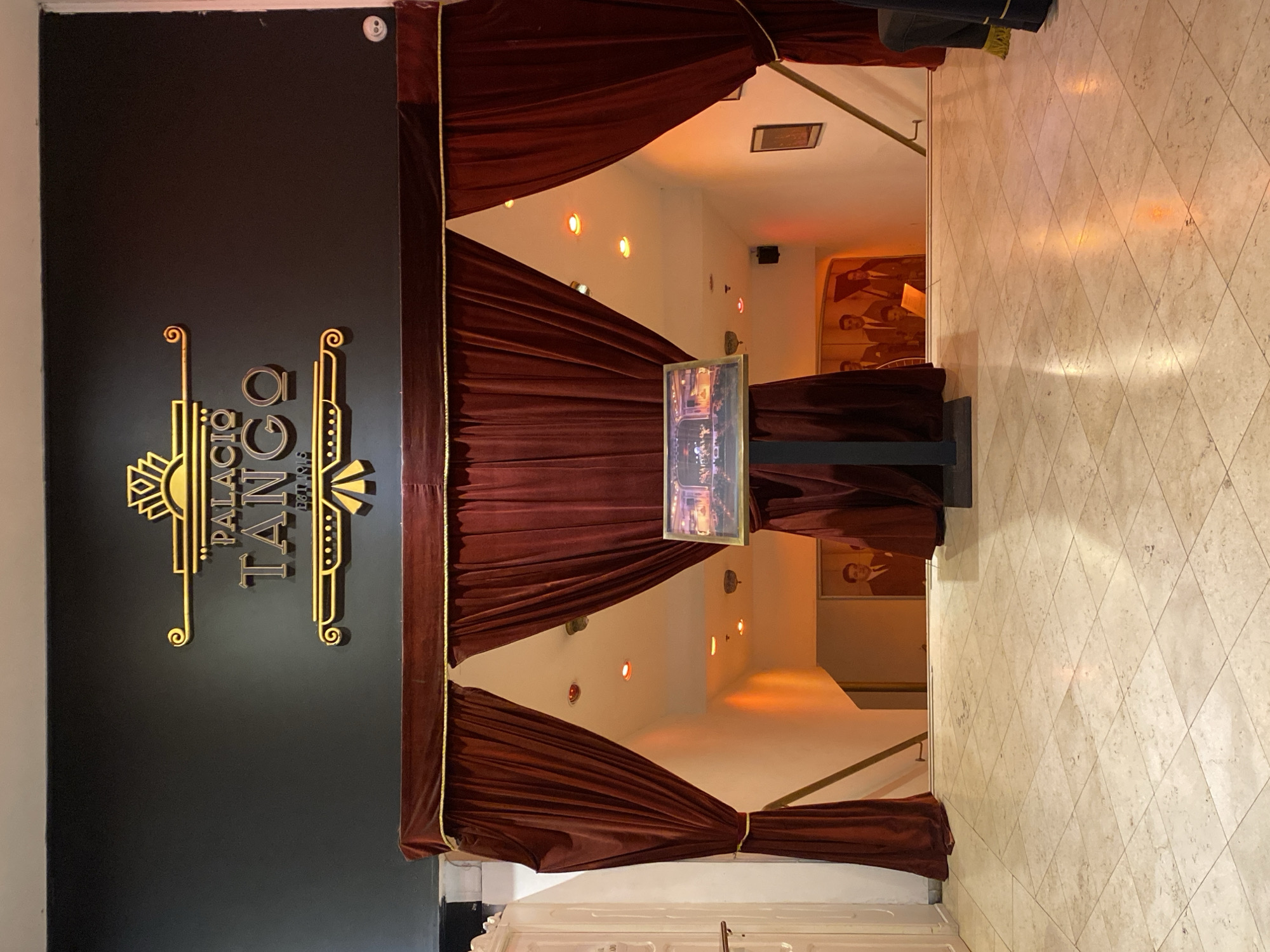 The image depicts the entrance to Palacio Tango in Buenos Aires, Argentina. A black wall on the left displays a golden art deco-style logo. A dark red curtain frames an entranceway, revealing an illuminated interior with white walls and red accent lights. Inside, a framed image sits on a stand, partially obscuring a mural of men in suits. The floor is covered in a tiled pattern. The scene appears devoid of people.