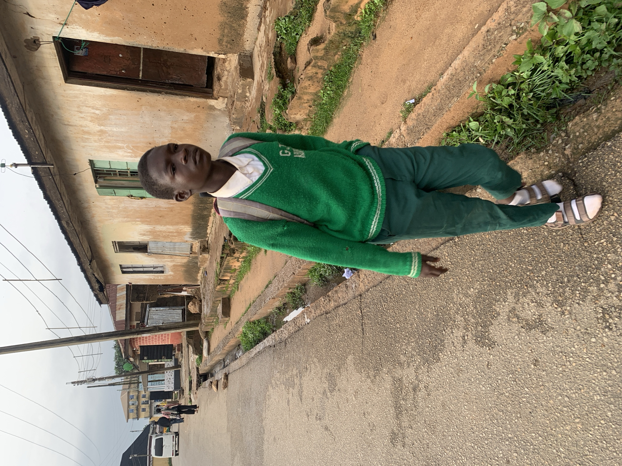 This media file captures a young boy in a school uniform, standing on a paved street in what appears to be a residential or mixed-use area. The boy is wearing a green V-neck sweater over a white collared shirt, and green trousers. He has a backpack slung over his right shoulder. His feet are clad in white socks and brown sandals.
The scene is set in a somewhat weathered urban environment. Behind the boy, there's a building with a light brown, textured wall, featuring a dark, rustic-looking door and a green-framed window. A narrow, overgrown ditch runs alongside the street, with patches of green vegetation pushing through the dirt and concrete. Further down the street, other buildings and structures are visible, including what seems to be shops or businesses with signage, and telephone poles with a network of wires. Some people can be seen in the distance, walking along the street. A white van is also parked on the side of the road.
The lighting suggests it could be daytime, possibly overcast given the muted quality of the light and the somewhat hazy sky in the background. The overall impression is of a typical street scene in a developing urban area. There is no discernible text visible in the image. The boy's expression is neutral.