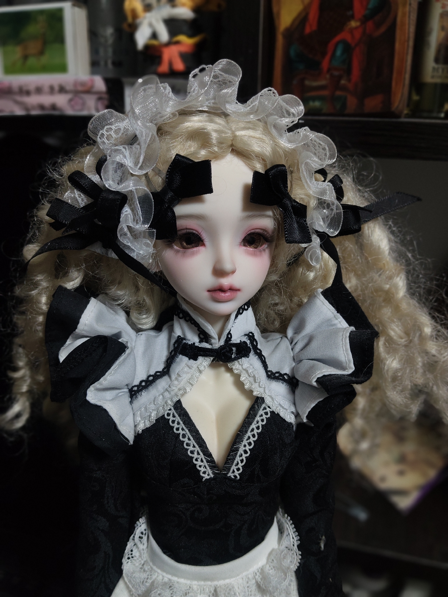 A close-up, eye-level, vertical shot shows a doll with blonde, curly hair, dressed in a black and white maid outfit. The doll has fair skin, pink cheeks, and dark brown eyes. It wears a white lace headband adorned with black bows. The outfit features a black lace bodice with a white ruffled collar and sleeves, and a white apron with lace trim. The background is dark and out of focus, with some shelves and objects visible, including a framed picture of a deer and what appears to be other toys or collectibles. The image is well-lit, highlighting the doll's features and attire. There is no discernible location in Kharkiv, Ukraine.