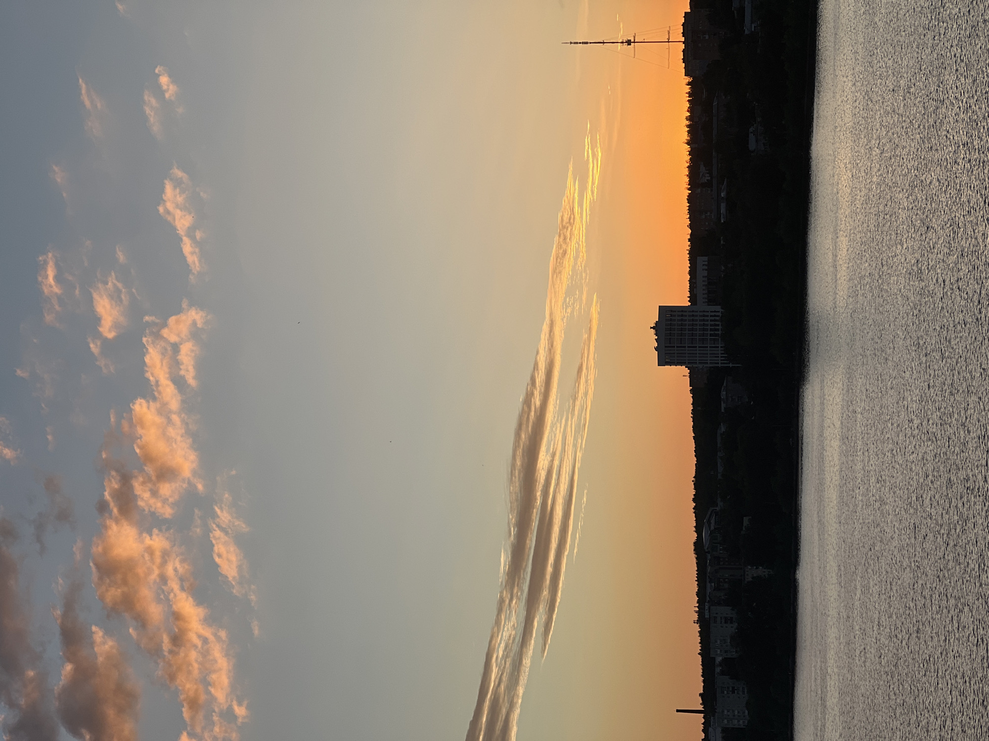 The image depicts a body of water in the foreground, with a silhouetted shoreline in the middle ground and a sunset sky in the background. On the shoreline, there are buildings and trees. A tall, thin antenna-like structure is visible on the right side of the image. The sky is a gradient of colors, transitioning from a light blue at the top to a vibrant orange near the horizon, with streaks of pink and orange clouds. The water's surface is rippled, reflecting the colors of the sky. The scene appears to be a serene evening view of a city, likely Izhevsk, Russia, given the presence of urban structures against a natural water body and sky.