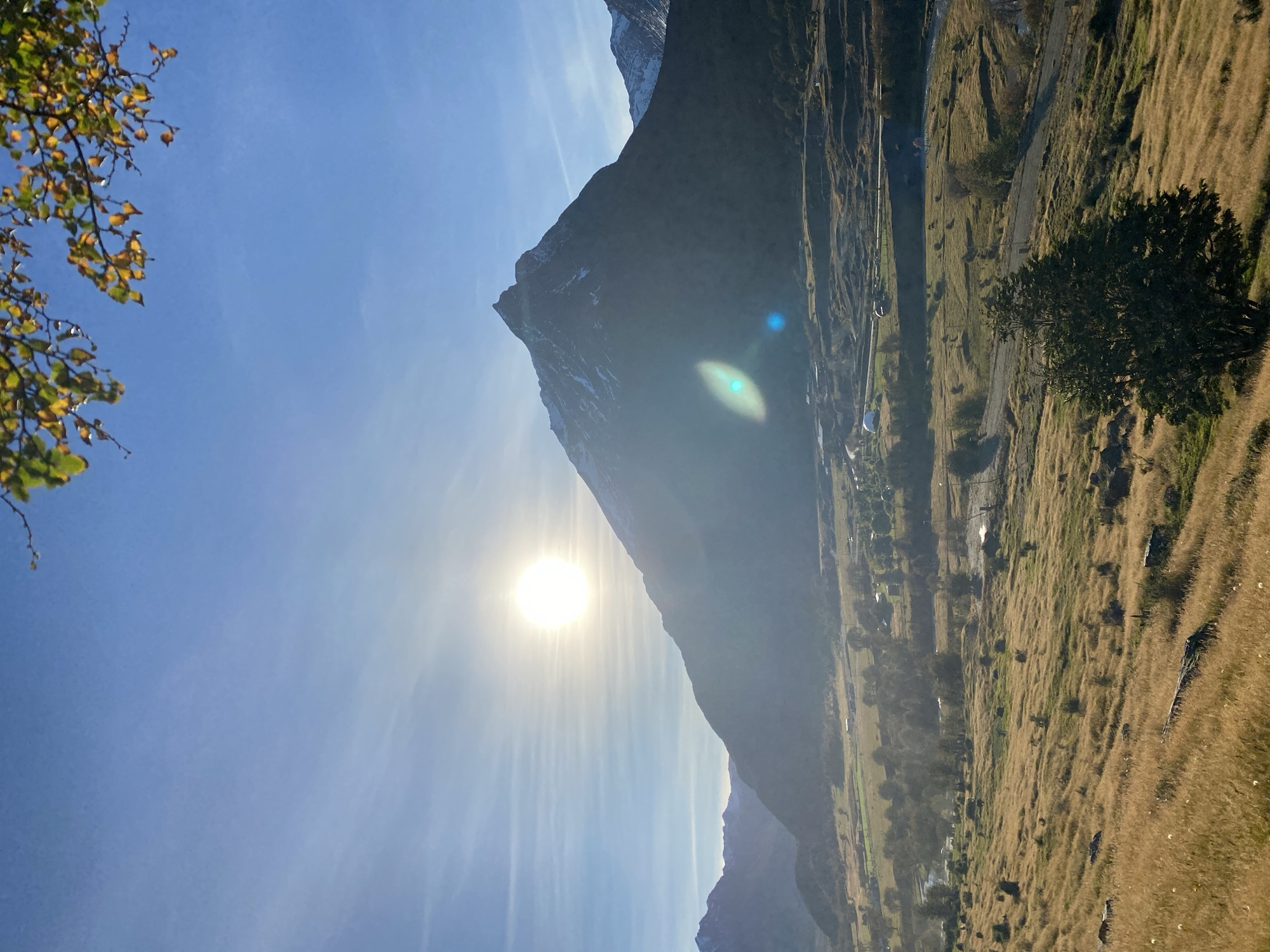 The image depicts a mountainous landscape in Ushuaia, Argentina. The sun is prominently positioned in the frame, radiating intense light and creating lens flare. A large, dark, triangular mountain dominates the scene, rising steeply from the valley floor. The foreground features a field of golden grass and a few trees. In the distance, there are indications of a road or path and scattered buildings or structures. The sky is clear blue with a few wisps of clouds. The scene captures the stark beauty of the Ushuaia landscape.