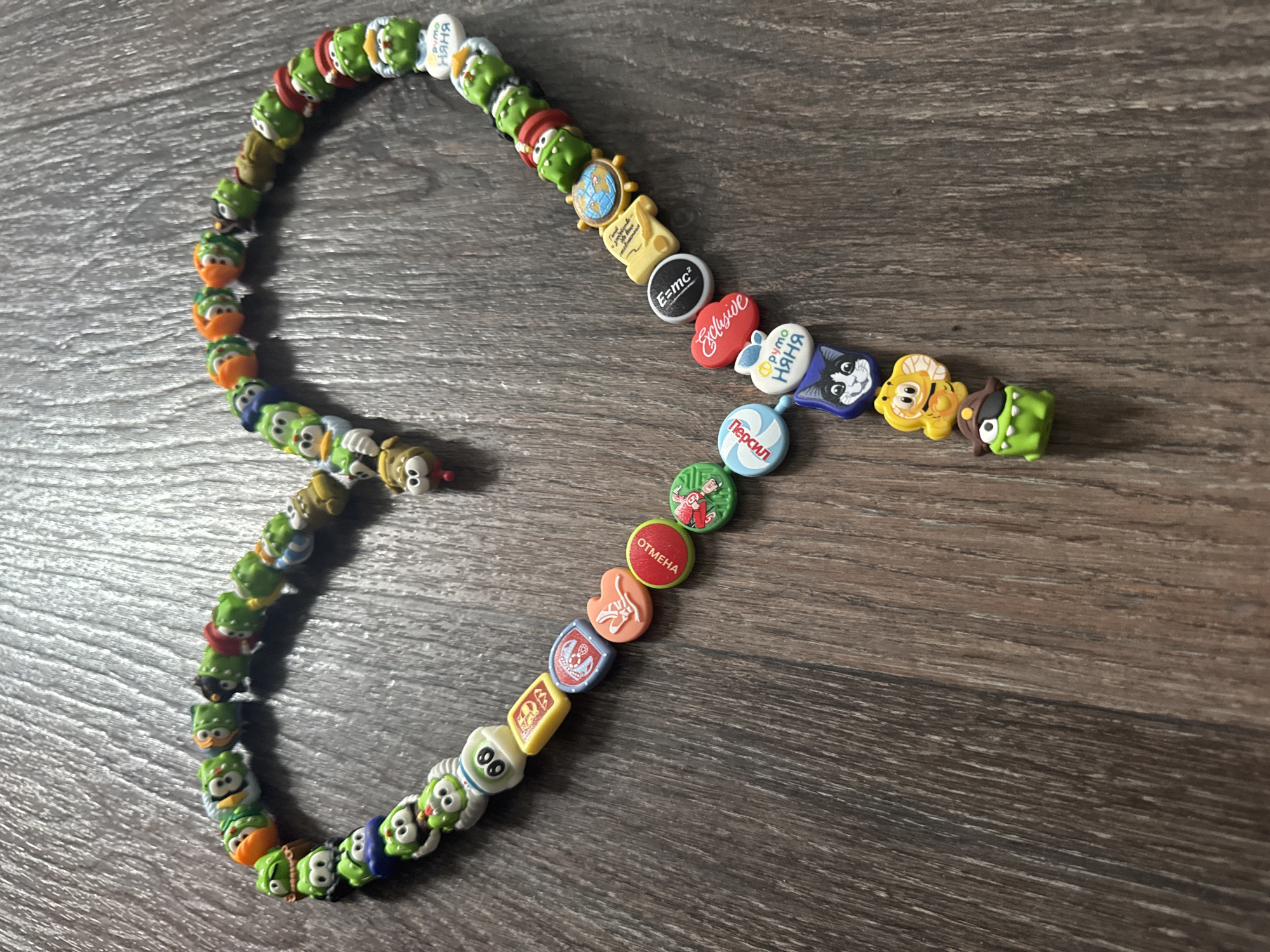 The image shows a collection of small, colorful plastic charms strung together, possibly forming a bracelet or necklace. Many of the charms depict cartoon characters, with a significant number featuring the "Smeshariki" characters. Other charms include miniature representations of objects like a globe, a book with the E=mc² equation, a red heart with the word "Exclusive," and various symbols and insignias. The charms are arranged on a dark wooden surface with a visible grain pattern. The location of the image is Chaikovsky, Russia.