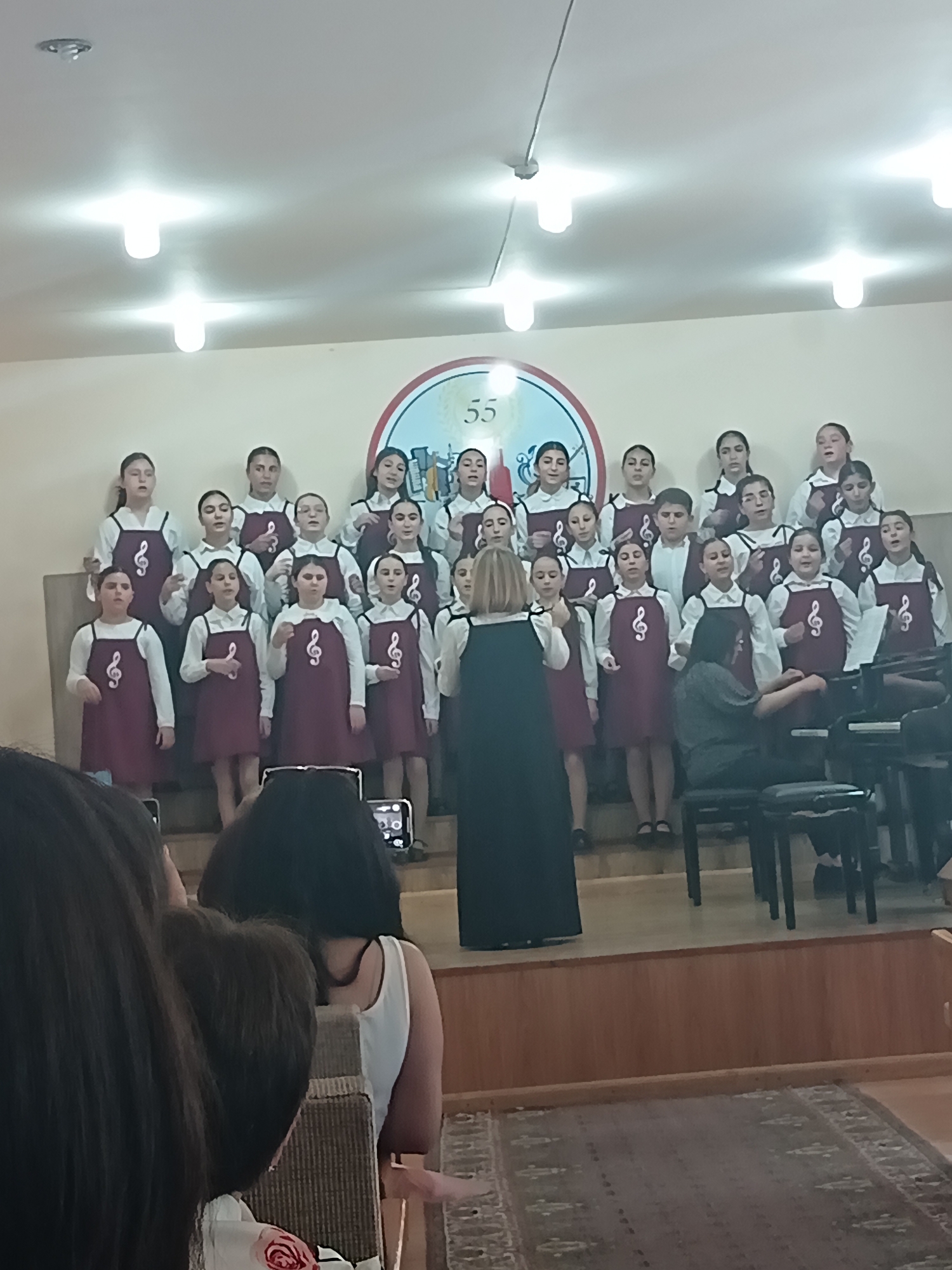 A group of young female choir members, dressed in white shirts and burgundy jumpers with treble clefs, stand on risers and perform on a stage. A woman in a black dress stands to the right of the choir, seemingly conducting or accompanying them, positioned near a grand piano. Another woman, partially visible in the foreground, is recording the performance with a phone. The background features a wall with a circular emblem displaying the number "55". The room appears to be an indoor venue, possibly a school or community hall, with stage lighting illuminating the scene.